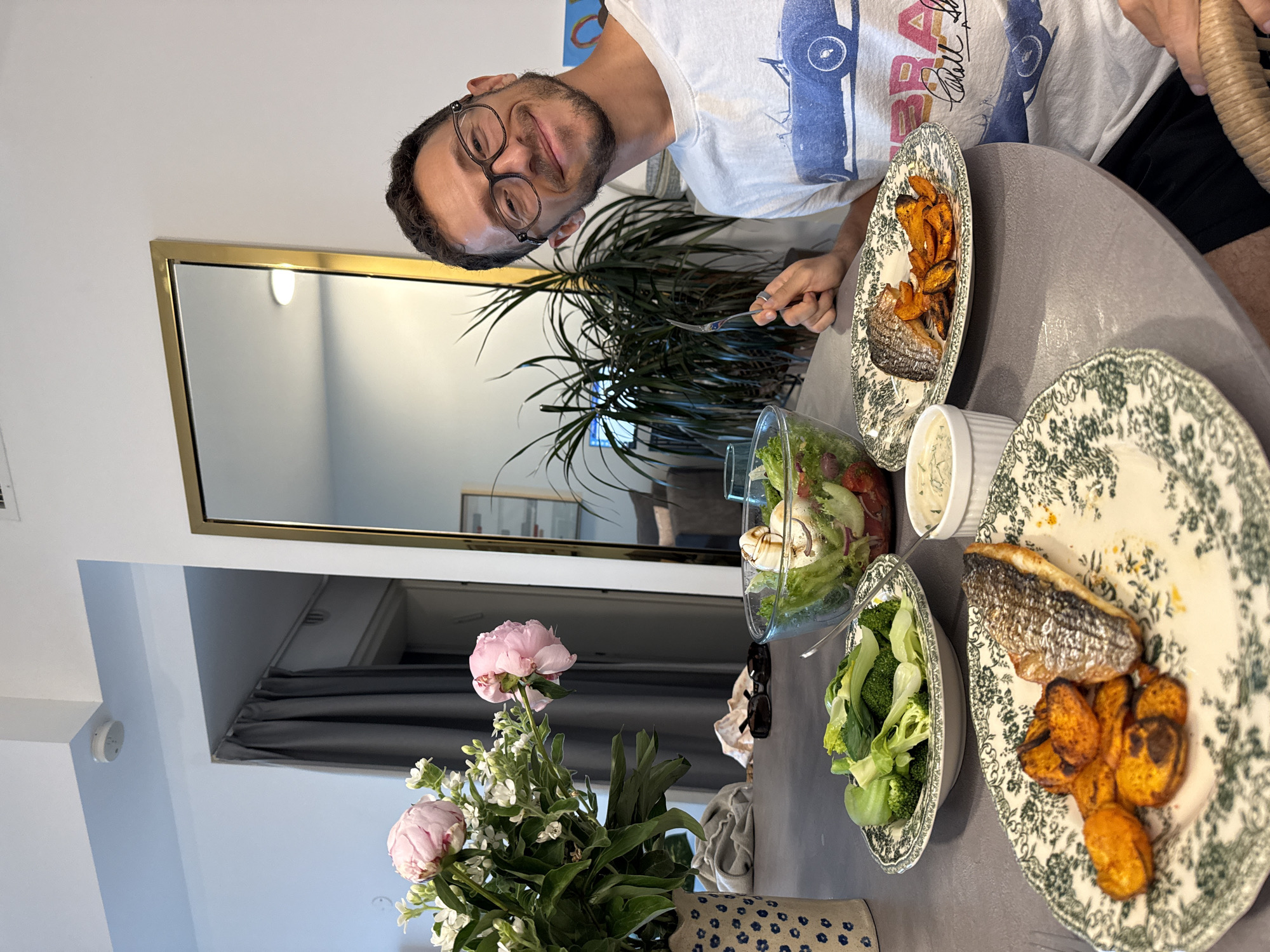 A man with glasses and a beard is seated at a table in what appears to be an apartment. The location could possibly be in Tel-Aviv, Israel, but that can not be determined from the image.
He has a plate with fish and sweet potatoes, a bowl of salad, and another of broccoli. A vase with pink flowers is also on the table. A mirror reflects the interior of the apartment.
