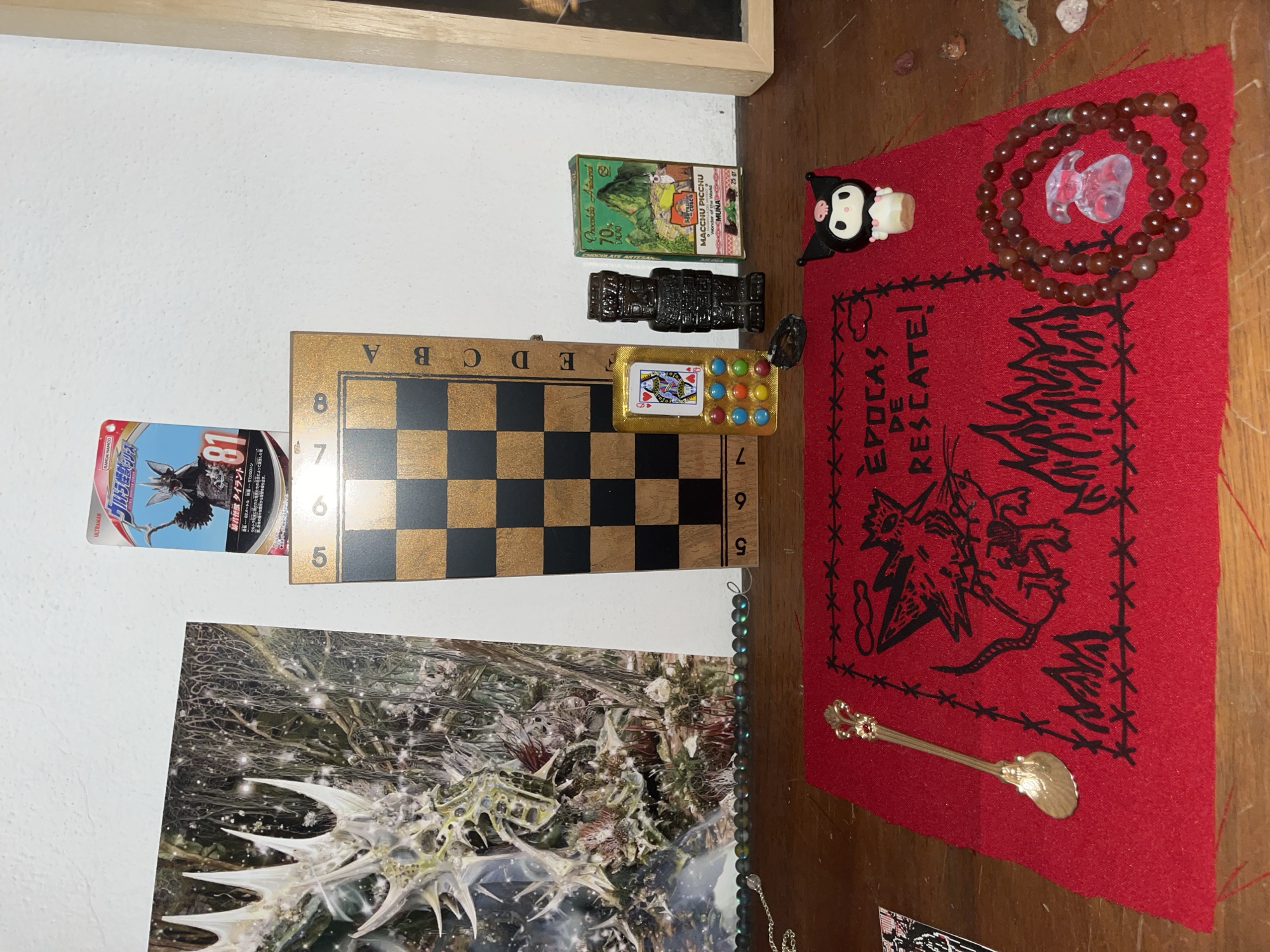 The scene presents a collection of diverse objects arranged on what appears to be a wooden surface. These items include a framed artwork, a small chessboard, a trading card, a miniature chocolate box labeled "MACCHU PICCHU", a dark figurine, a pill-shaped container with a playing card and colorful pills, a red cloth with printed designs and text that reads "ÉPOCAS DE RESCATE!", a bracelet made of brown beads, a clear glass charm, a small Kuromi figure, a small golden spoon, and beaded necklaces of varying colors. The image is rotated, with the surface oriented vertically, and the objects appear to be randomly scattered.