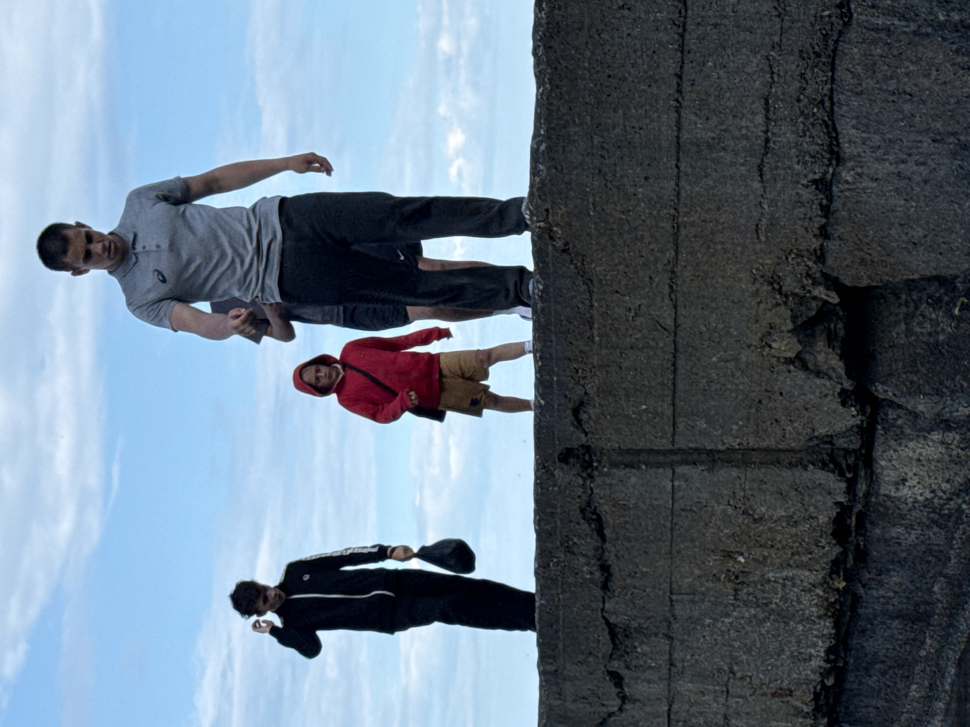 A group of four individuals, appearing to be juveniles or young adults, stand on a weathered concrete structure against a pale blue sky with scattered clouds. In the foreground, a person wearing a grey polo shirt and dark pants walks forward, looking down. Behind them and slightly to the left, a person in a red hooded jacket and light-colored shorts stands facing left. To the left of the person in red, a person in a black tracksuit with white stripes walks forward, carrying a dark bag in their right hand and reaching up to their hair with their left. Further to the left and behind the person in the tracksuit, another individual is partially visible, wearing dark clothing. The concrete structure appears to be a wall or embankment, showing signs of age and damage with cracks and uneven surfaces.