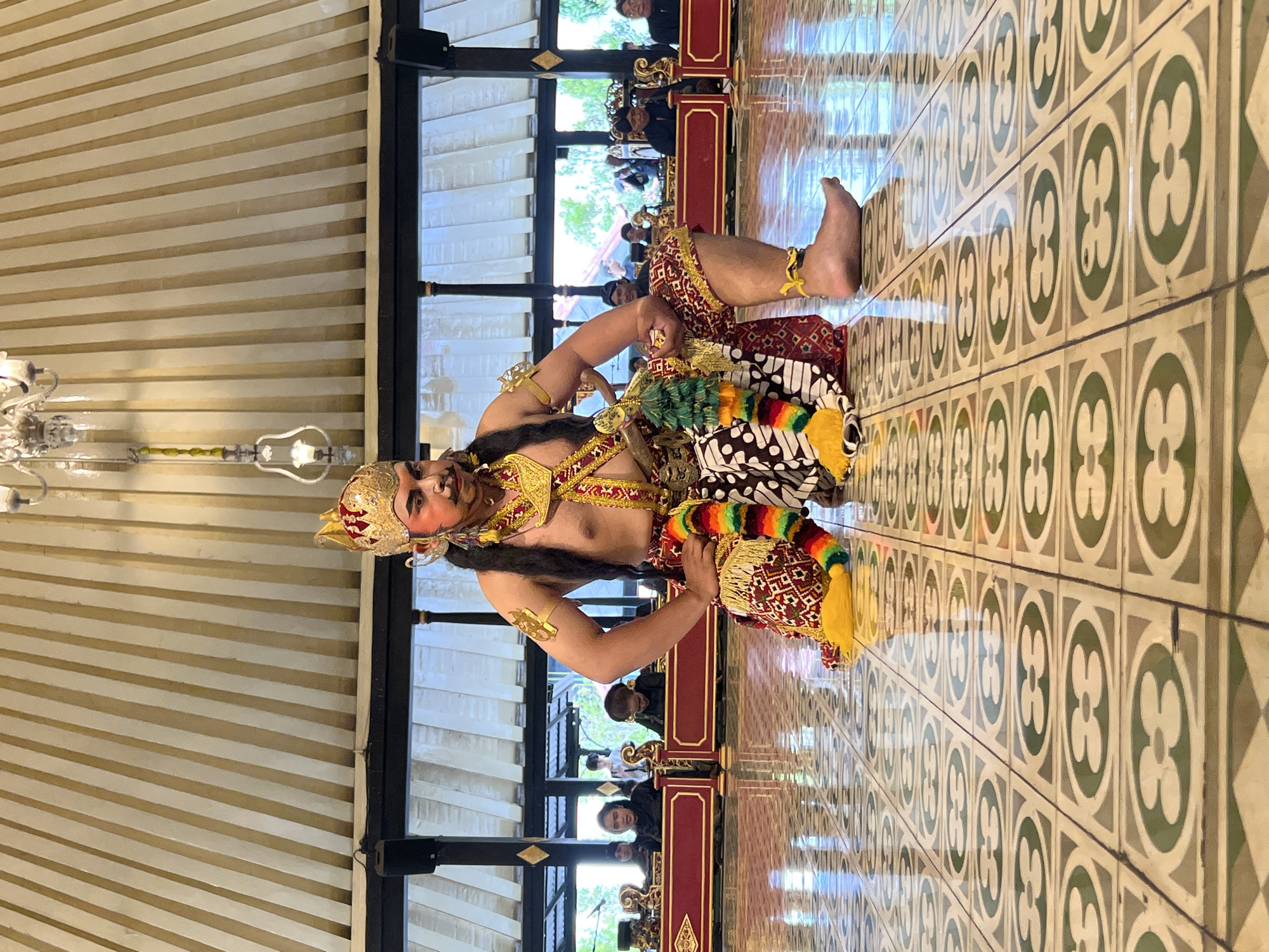 A male dancer in traditional Javanese attire kneels on a patterned tiled floor in a pavilion with a sloped, striped ceiling and ornate chandeliers. The dancer wears a gold and red headdress, elaborate gold jewelry, and a vibrantly colored sash. His face is painted with dramatic makeup. In the background, an ensemble of musicians sits behind a red wooden barrier, playing traditional Indonesian instruments. The scene is set in the Kraton (Sultan's Palace) in Yogyakarta, Indonesia, a location renowned for preserving Javanese culture and arts. The pavilion architecture and the presence of the gamelan orchestra indicate a cultural performance, likely a traditional dance or theatrical presentation within the palace grounds. The polished floor reflects the overhead lights and the figures in the scene, creating a sense of depth and grandeur. The audience members in the background are partially visible, suggesting an ongoing event.