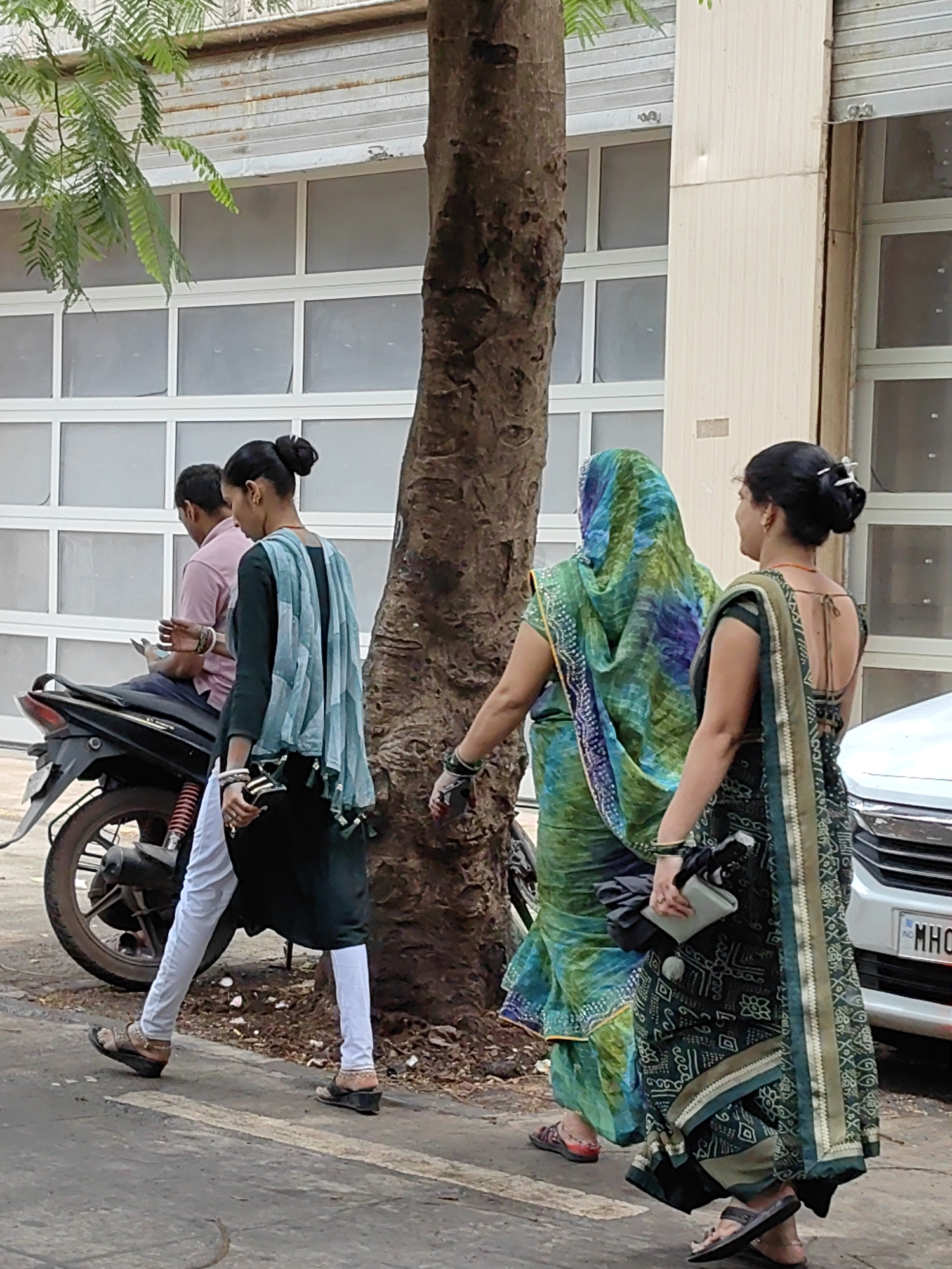 In Mumbai, India, three individuals are observed walking beside a tree on a street, with a motorcycle parked to the left and a white car partially visible to the right. The first individual, a man wearing a pink shirt and blue trousers, is seated on the motorcycle and appears to be looking at his phone. Beside him, a woman dressed in a dark green kurta and light blue dupatta walks with her back partially turned, her hair styled in a bun. Further to the right, another woman, wearing a green and blue patterned sari, walks ahead. A third woman, in a dark green sari with an intricate pattern, is walking in the foreground, holding a grey purse. The background features a large, roll-up garage door and a section of building facade. The ground is paved, with a faint crosswalk marking visible.