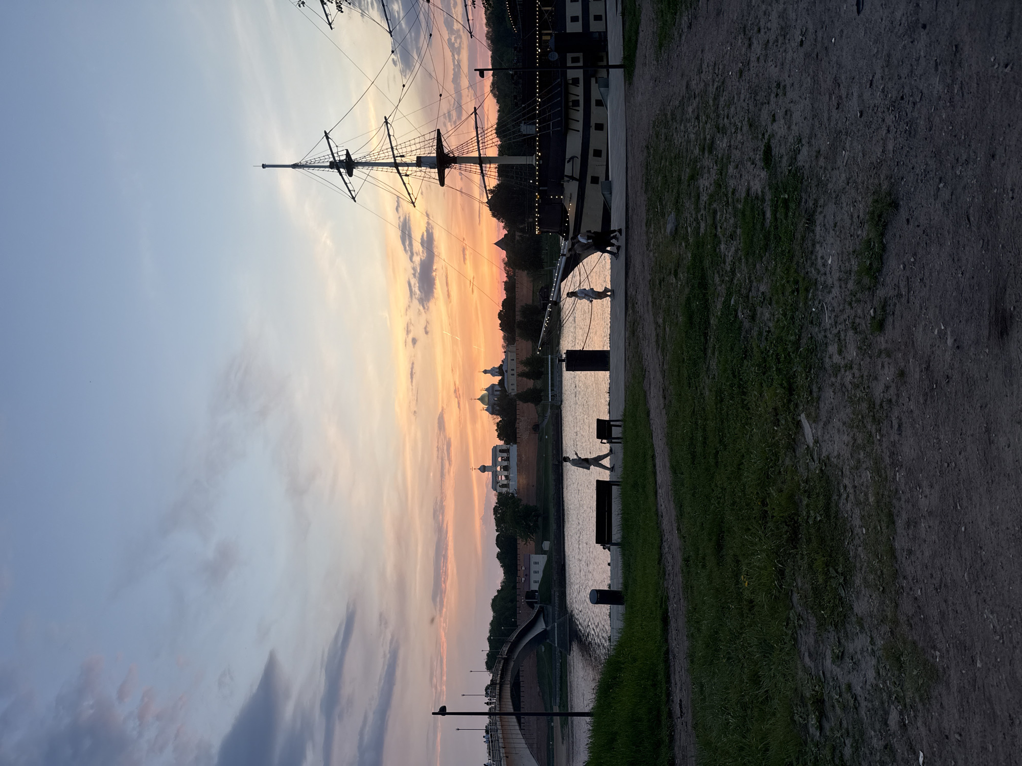 The image depicts a twilight scene in Veliky Novgorod, Russia. On the left is a pastel sky with shades of orange and pink near the horizon, transitioning to a bluish-grey higher up. To the right of the sky is the silhouette of a tall ship's rigging and mast. The body of the ship appears dark with some lights visible.
Below the ship, across the river, are the domes of orthodox churches and a fortified wall, likely the Kremlin. The river reflects the colors of the sky and the silhouettes of people walking along the embankment, along with benches and trash receptacles.
In the foreground, the right side of the image, is a path made of compacted dirt and gravel. Adjacent to the path is a strip of grass, indicating a park-like setting.