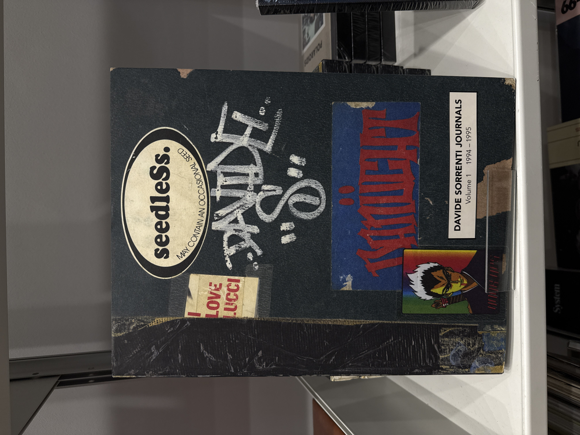 A single, stylized journal titled "DAVIDE SORRENTI JOURNALS Volume 1 1994 - 1995" rests on a white shelf, likely within a bookstore, gallery, or exhibition space in New York City, United States of America. The journal features a dark, textured cover with a collage of stickers and graffiti. An oval white sticker at the top center displays "seedleSs." in black and "MAY CONTAIN AN OCCASIONAL SEED." Below this, white graffiti-style text reads "DAVIDE "S"." To the upper left, a rectangular beige sticker states "I LOVE LUCCI" in red. A blue rectangular sticker with red graffiti-style text, possibly "IKNIGHT," is adhered below the white graffiti. In the bottom left corner, a small rectangular sticker depicts an anime-style character with white spiky hair, yellow eyes, and red markings, appearing to hold a sword. A white label at the bottom right identifies the journal as "DAVIDE SORRENTI JOURNALS Volume 1 1994 - 1995." The journal's left edge and corners show signs of wear or intentional distress. To the right of the journal, a stack of black, shrink-wrapped items, possibly books or media, is visible, with "POLAROIDS" partially discernible on one spine. A grey shelf is positioned above and to the left, while another shelf with partially visible items, including one labeled "System," is below.