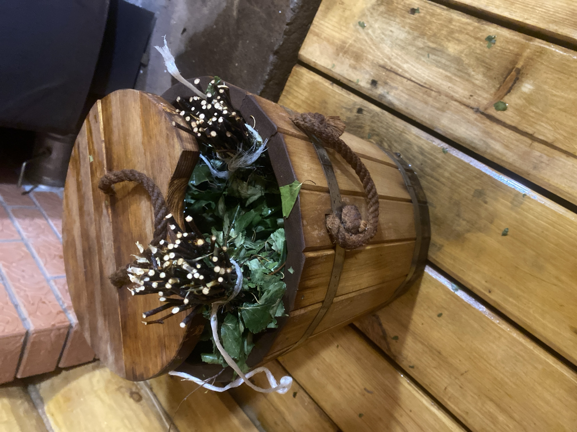 The image depicts a wooden bucket filled with leafy branches and decorative twigs. The bucket is made of light brown wood, with darker brown bands securing the staves together. A thick rope handle is attached to the sides of the bucket. Inside, there are green leafy branches, and bundles of twigs decorated with small, white, round objects. The bucket is placed on a surface made of wooden planks. Part of a brick floor is visible to the left. The location is Гаревая, Russia. There are no people visible in the image. The environment is indoors. The objects are a wooden bucket, leafy branches, and decorative twigs. There are no actions or interactions visible in the image.