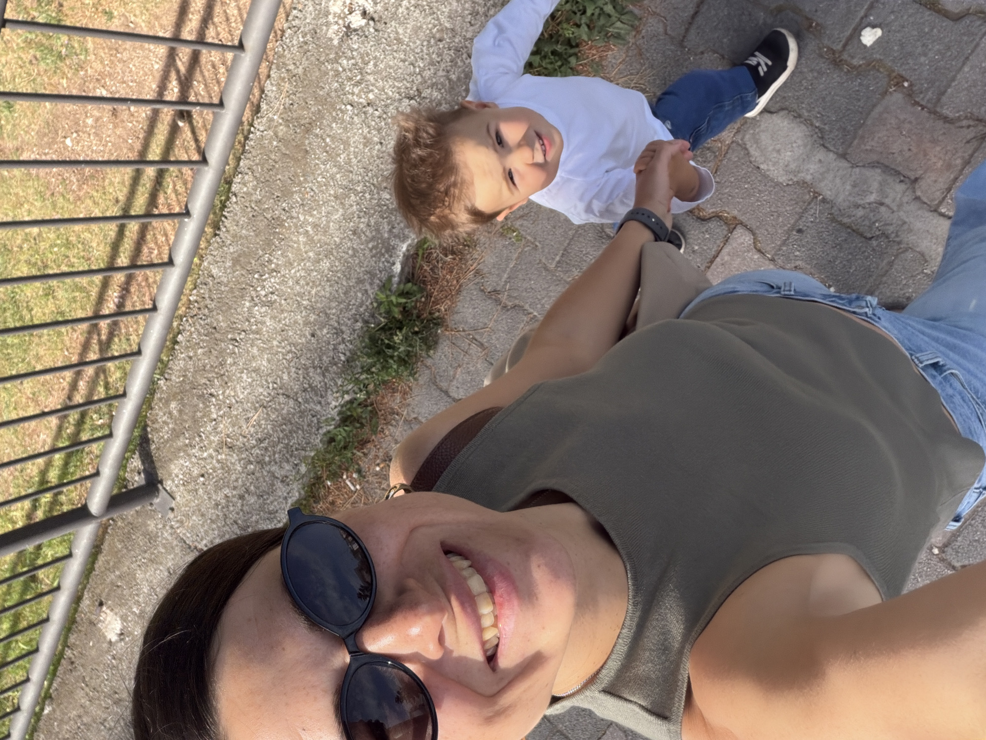 A selfie-style low-angle full shot shows a woman and a young child on what appears to be a paved walkway. The woman, wearing sunglasses and a gray tank top with a shoulder bag, is smiling broadly. The child, dressed in a white long-sleeved shirt and blue jeans, is looking upwards and holding the woman's hand. The background includes a metal railing, a sidewalk, and sparse greenery. The location could be in Budva, Montenegro, near an area with pedestrian walkways and railings, perhaps along the coast or in a park-like setting.
