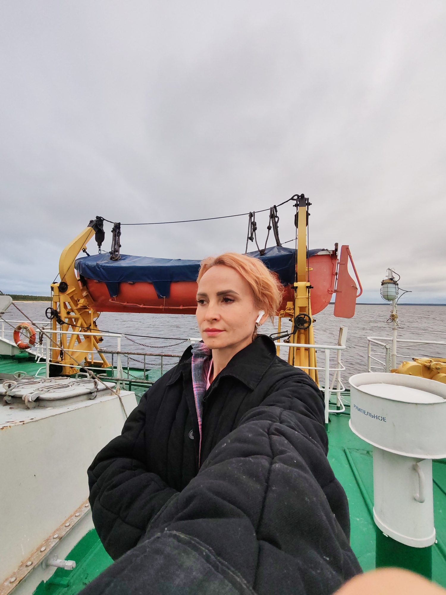 A woman with short, reddish-blonde hair is taking a selfie on the deck of a ship. She is wearing a black quilted jacket and appears to have white wireless earbuds in her ears. Behind her, a life raft is suspended from a yellow davit. The ship's deck is green, and a white railing runs along the side. In the background, a body of water stretches to the horizon under a cloudy sky. The location is identified as Ozernoe Ust'e, Russia, a port city located on Lake Baikal.