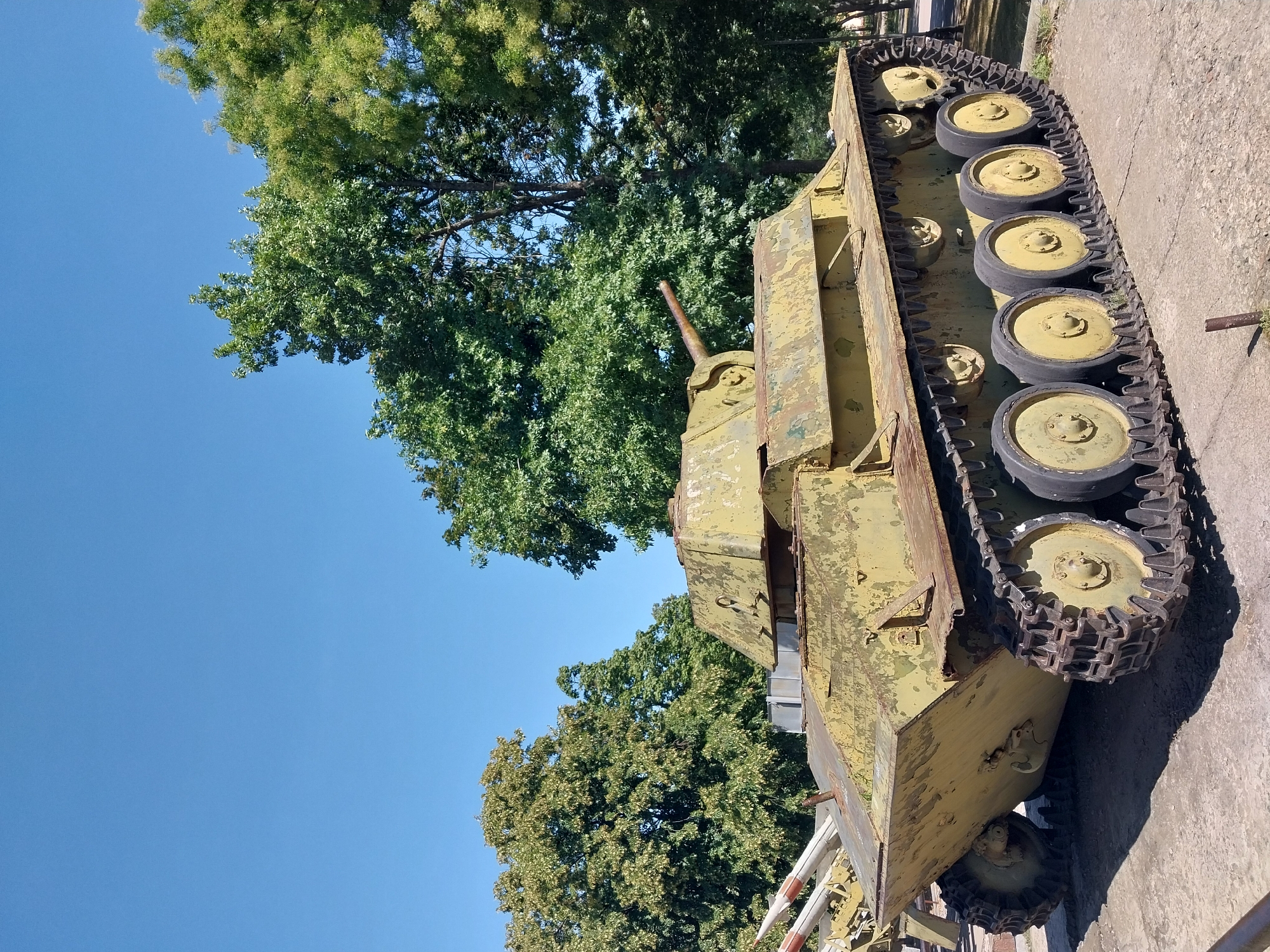 A weathered, light green tank sits on a concrete surface under a clear blue sky, with lush green trees in the background. The tank, an older model with visible rust and peeling paint, has a turret with a visible gun barrel. To the left of the tank, parts of a rocket launcher are visible. The scene appears to be an outdoor exhibit, possibly a memorial or museum, in Dnipro, Ukraine. There are no people present. The ground is a paved surface with a few small metal posts. The lighting suggests it is daytime, with shadows cast by the tank and trees.