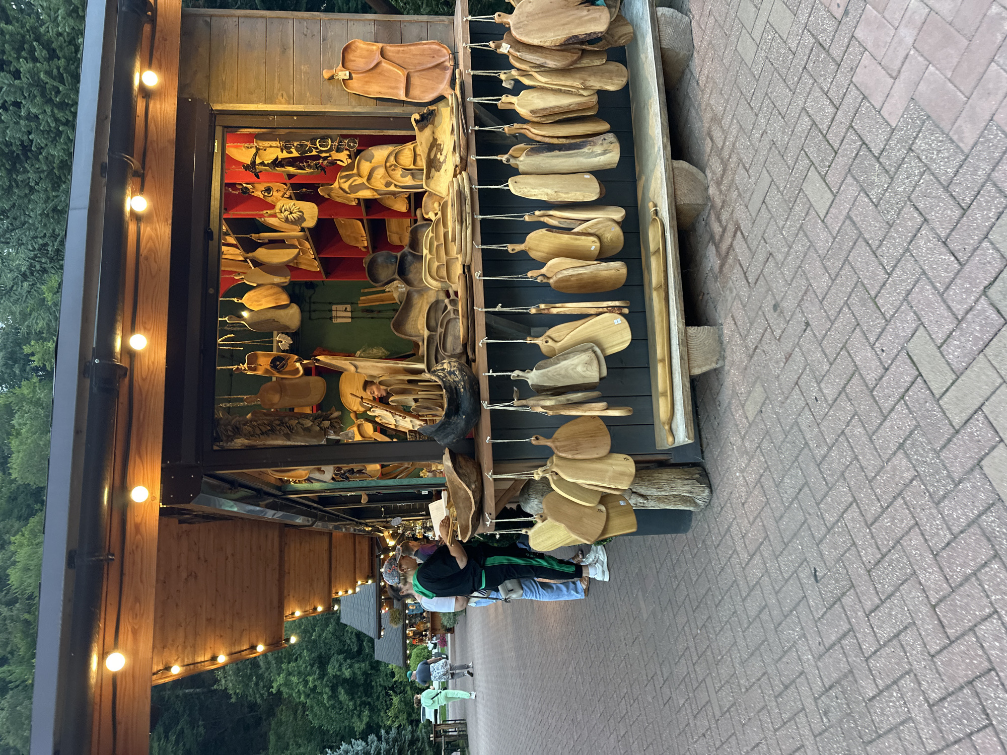 In Estosadok, Russia, a market stall displays an array of wooden kitchenware, primarily cutting boards and serving platters. The stall, constructed with wood, is illuminated by string lights overhead. The items are hung from the stall's front or displayed on shelves behind a counter. A few individuals are seen browsing the merchandise. The ground in front of the stall is paved with brick. The background reveals more market stalls and lush green trees.