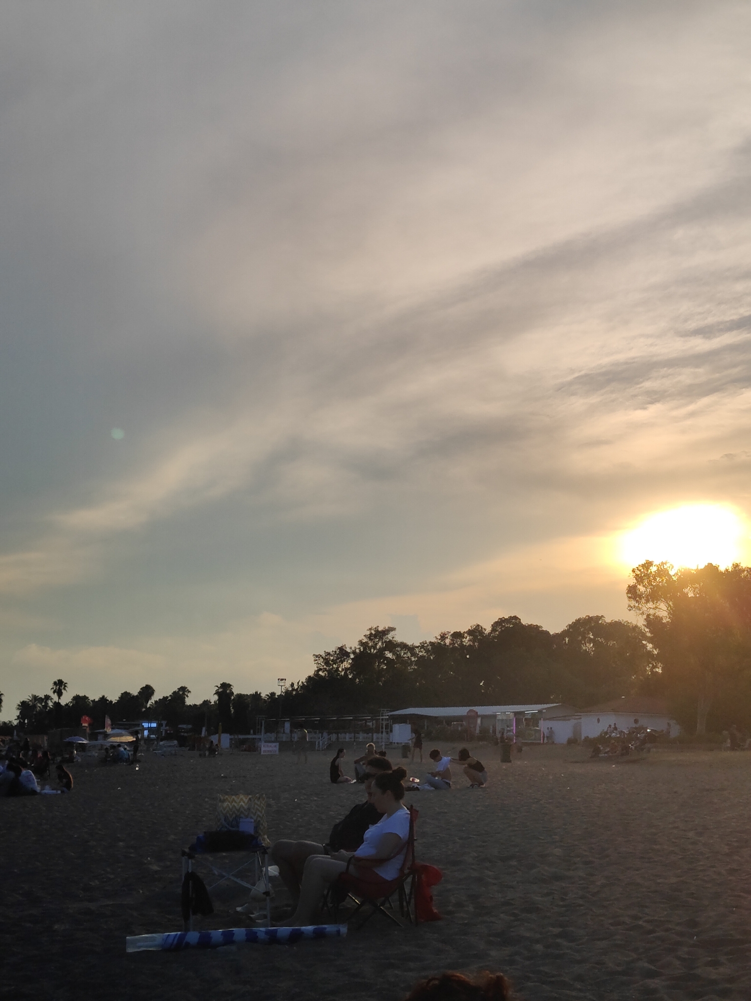 The image depicts a crowded beach scene during sunset in Antalya, Turkey. The sky is filled with soft, diffused sunlight and light clouds, with the sun itself a bright, glowing orb peeking through the trees on the right side of the frame. The beach is covered in dark sand, and a line of trees with palm trees interspersed forms the background. Several small buildings, possibly shops or beachside facilities, are visible behind the trees. Numerous people are scattered across the sand, some sitting in chairs, others gathered in groups, and a few are walking. In the foreground, two individuals are seated in camping chairs facing away from the camera, with a small table and a rolled-up mat on the sand between them. The overall atmosphere appears relaxed and social, characteristic of a popular beach destination during the evening hours.