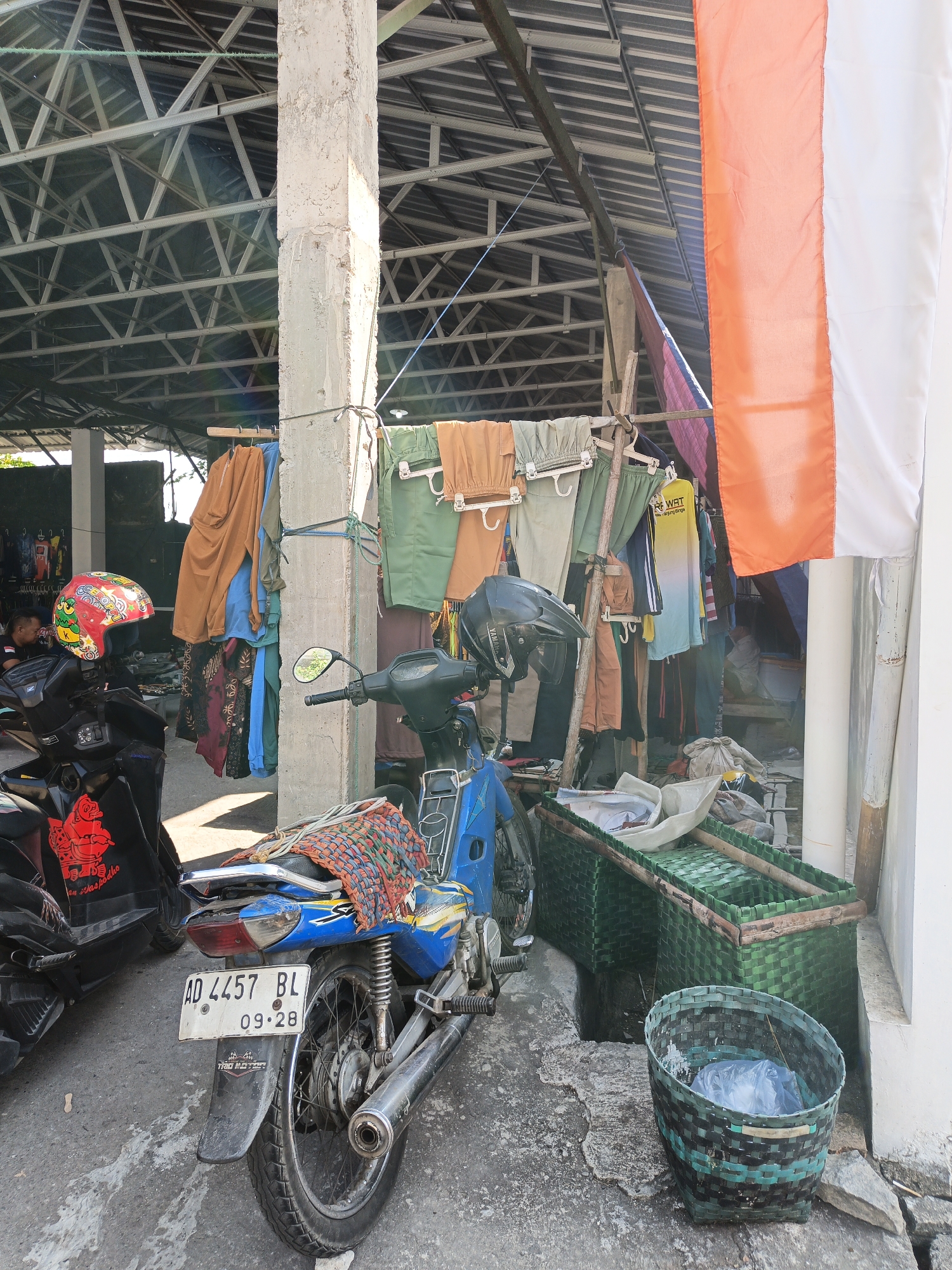 The image depicts an outdoor scene in Jatinom, Indonesia, likely a market or roadside stall. A concrete pillar supports a corrugated metal roof structure. Several motorcycles are parked, including a blue one with a license plate reading "AD 4457 BL 09-28." A red and yellow helmet rests on the handlebars of a black motorcycle. Clothes are hung out to dry on a rack made of bamboo poles and clotheslines, with various garments including pants and shirts in different colors. A large Indonesian flag, featuring vertical stripes of red and white, is displayed on the right side of the frame. In the foreground, there are two woven green baskets, one containing plastic bags, and a portion of a drain or ditch. The ground is paved with concrete.
