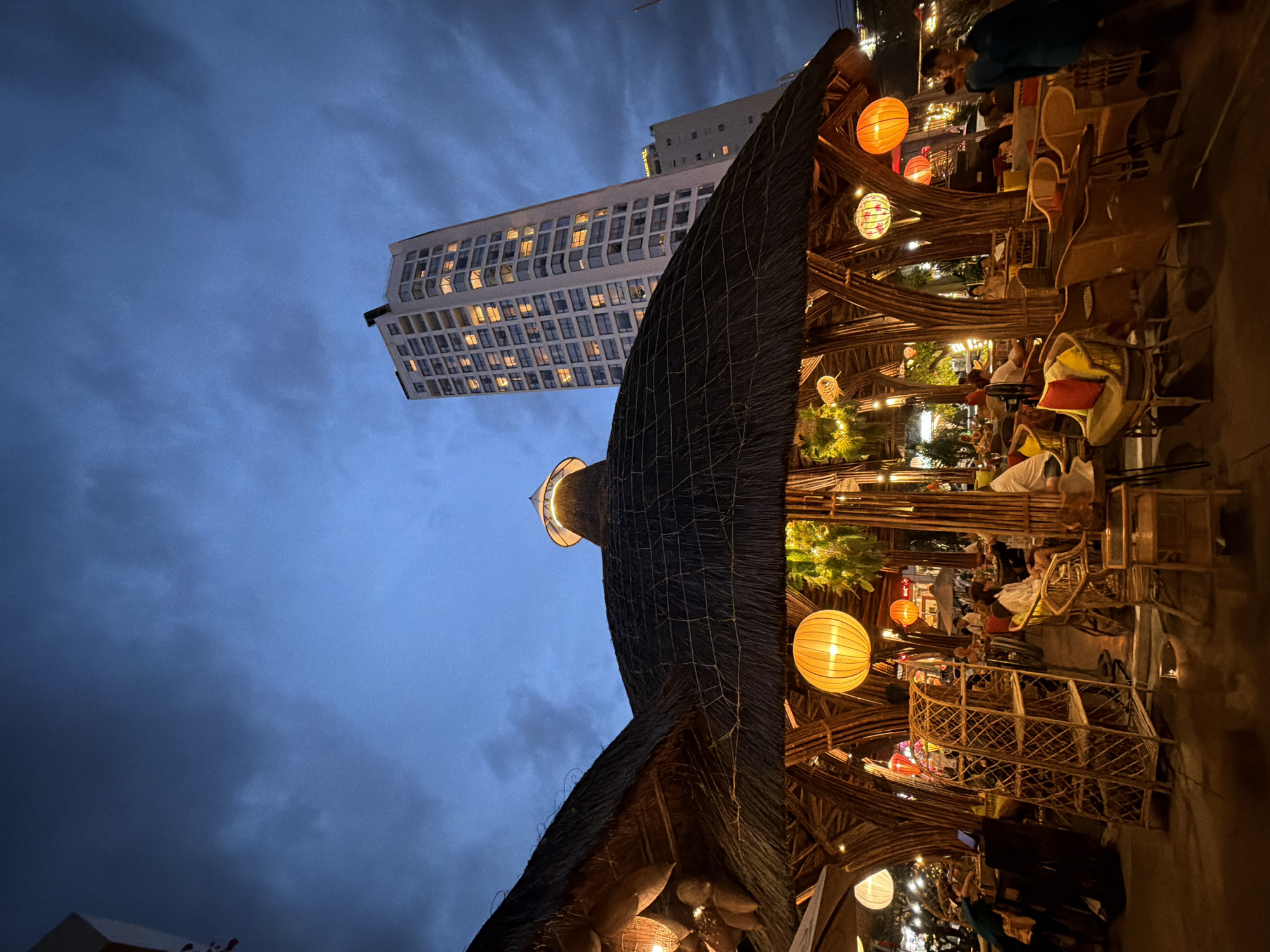 The photograph captures an exterior view of a restaurant with a thatched roof, featuring bamboo structures and outdoor seating. Large, illuminated paper lanterns hang from the ceiling. Several patrons are seated at tables, some appearing to be dining. In the background, a tall, modern high-rise building with illuminated windows is visible against a darkening sky with visible clouds. The scene suggests a nighttime setting, likely in a bustling urban area. The location is specified as Nha Trang, Vietnam.