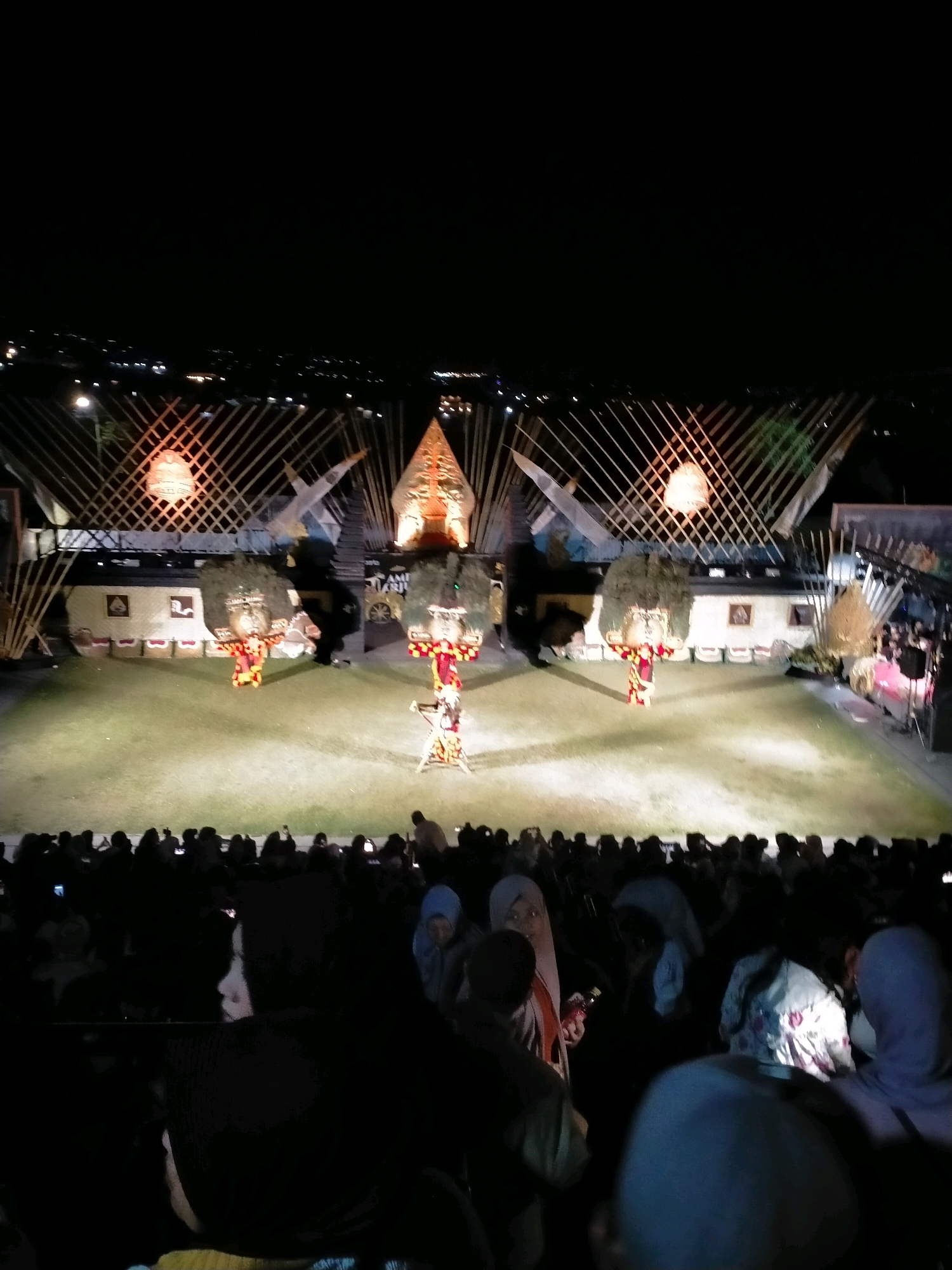 An outdoor night performance is taking place on a grass stage in front of a seated audience. The stage is decorated with bamboo structures and traditional Indonesian puppets. Two performers in colorful costumes are on stage, one in the center and another to the right. The audience is a crowd of people of various ages and ethnicities, some of whom are holding up phones to record the event. The backdrop of the stage features a cityscape with lights, suggesting the location is the city of Batu, Indonesia.