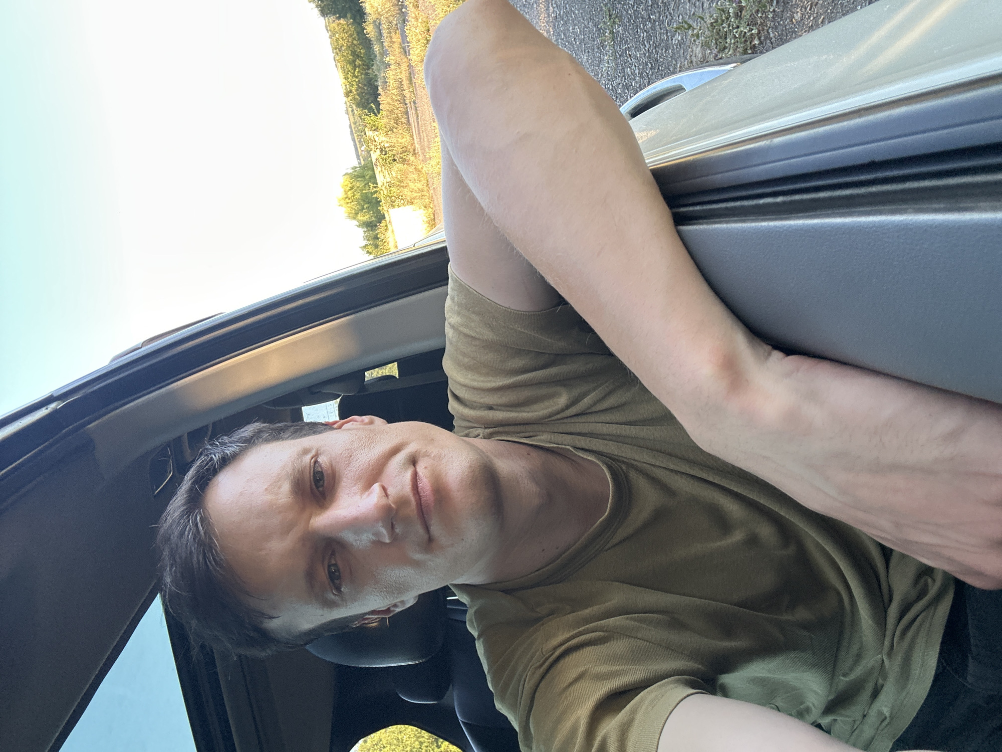 A man is sitting in the driver's seat of a car, with his arm resting on the open door frame. He is looking directly at the camera with a neutral expression. The car's sunroof is open, revealing a clear sky. The background shows a rural landscape with trees and vegetation, indicative of a location outside of a city. The lighting suggests it is daytime, likely late afternoon or early evening. The overall setting is informal and relaxed.