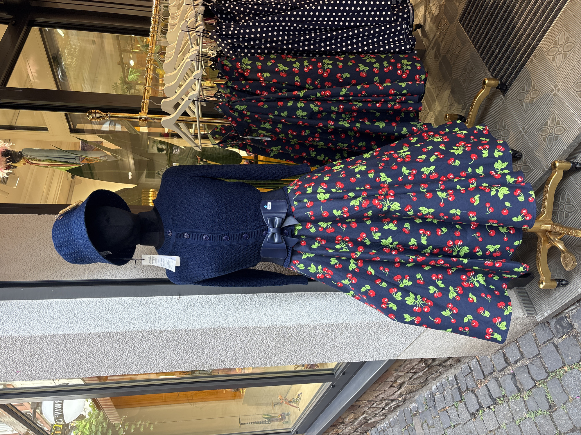 A mannequin dressed in a dark blue cardigan, a dark blue belt with a bow, and a dark blue skirt with a red cherry pattern stands outside a shop in Rüdesheim, Germany. The mannequin wears a dark blue hat. To the right of the mannequin, clothes on hangers are displayed on a gold rack, including dark blue skirts with red cherry patterns and a dark blue skirt with white polka dots. The shop entrance features a large window with reflections of the interior. The ground outside the shop is paved with cobblestones.