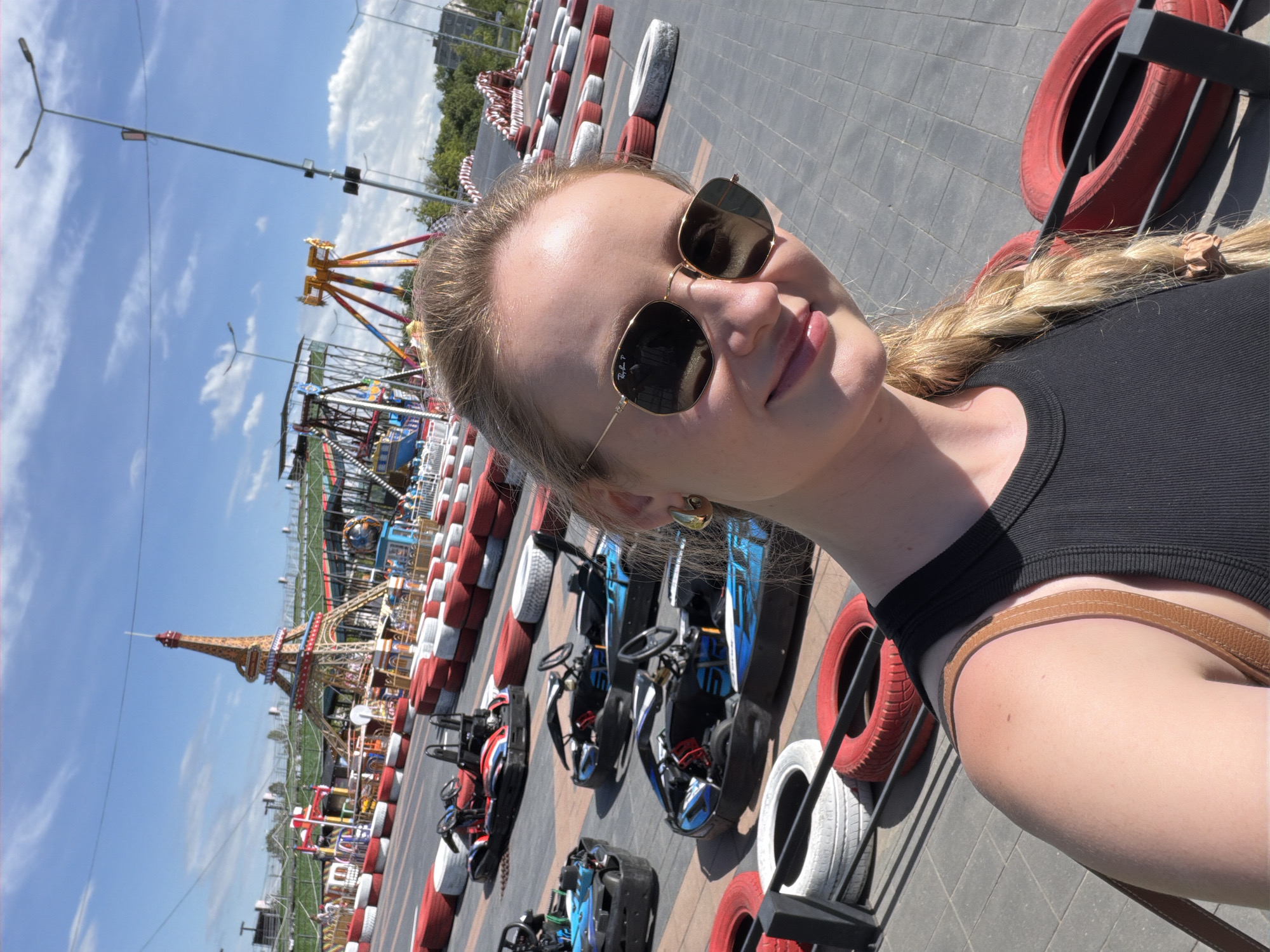 In Stăuceni, Moldova, a young woman with blonde hair braided down her back smiles at the camera, wearing sunglasses and a black tank top. She is positioned in the foreground of an outdoor go-kart track. The track is lined with red and white tires, and several go-karts are visible on the asphalt. In the background, amusement park rides are present, including a structure resembling the Eiffel Tower, a swing ride, and other attractions. The sky is blue with scattered clouds, and the scene appears to be a sunny day at an amusement park or entertainment complex.