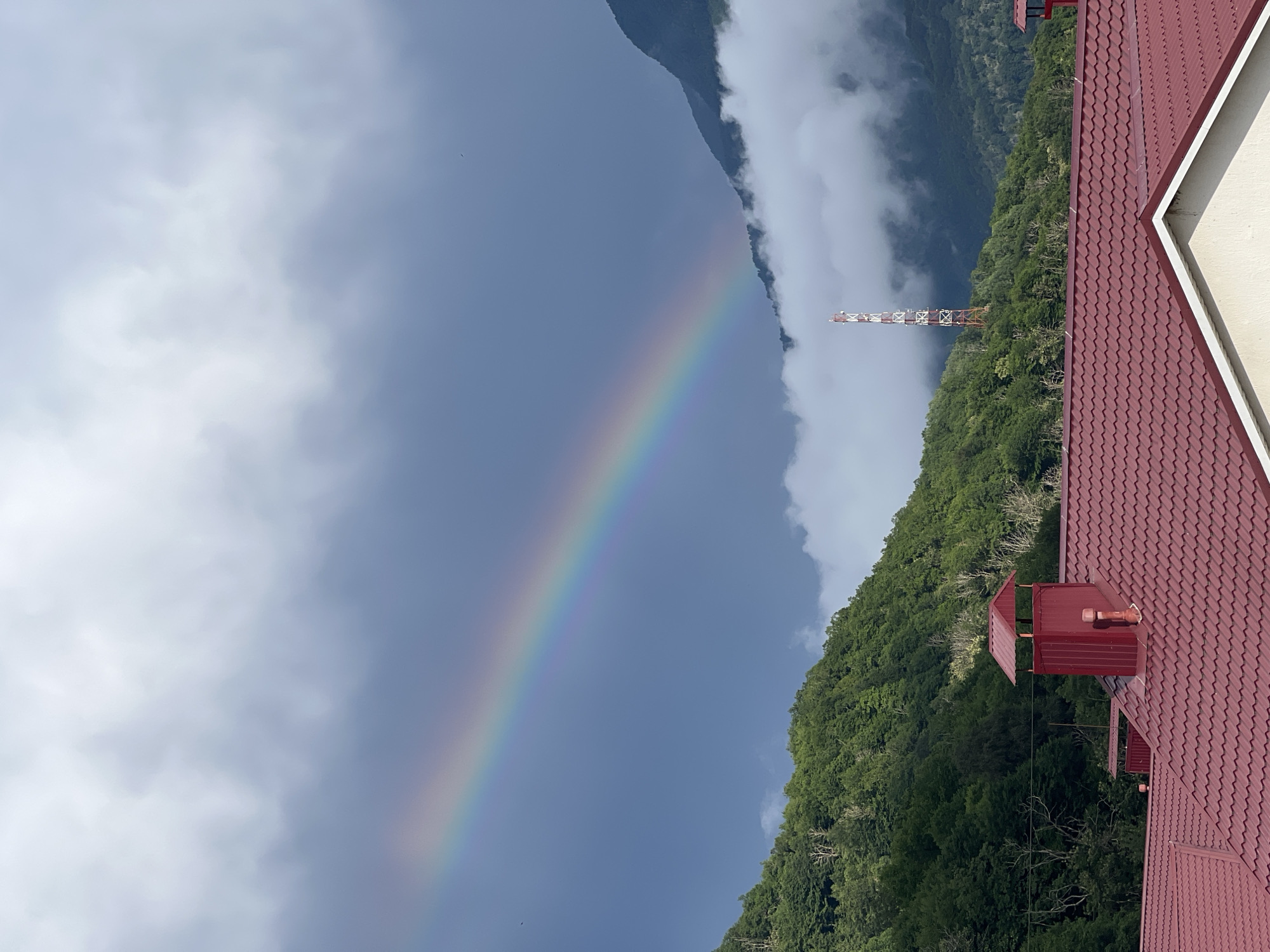 The image depicts a scenic mountain landscape with a rainbow arcing across a cloudy sky. The foreground features a portion of a red tiled roof, suggesting a building or structure situated on a mountainside. A small, red chimney or vent structure is visible on the roof. Lush green trees cover the slopes of the mountain, which are partially obscured by soft, white clouds. In the distance, a red and white lattice tower, likely a telecommunications or observation tower, stands on the mountainside, adding a man-made element to the natural scene. The presence of the rainbow suggests recent rainfall or ongoing precipitation in the area, typical of the weather in Krasnaya Polyana, Russia. The lighting and cloud cover imply it might be late afternoon or early morning, after a rain shower. The overall mood is tranquil and awe-inspiring, capturing the beauty of nature and the effect of weather phenomena. There is no visible text in the image.
