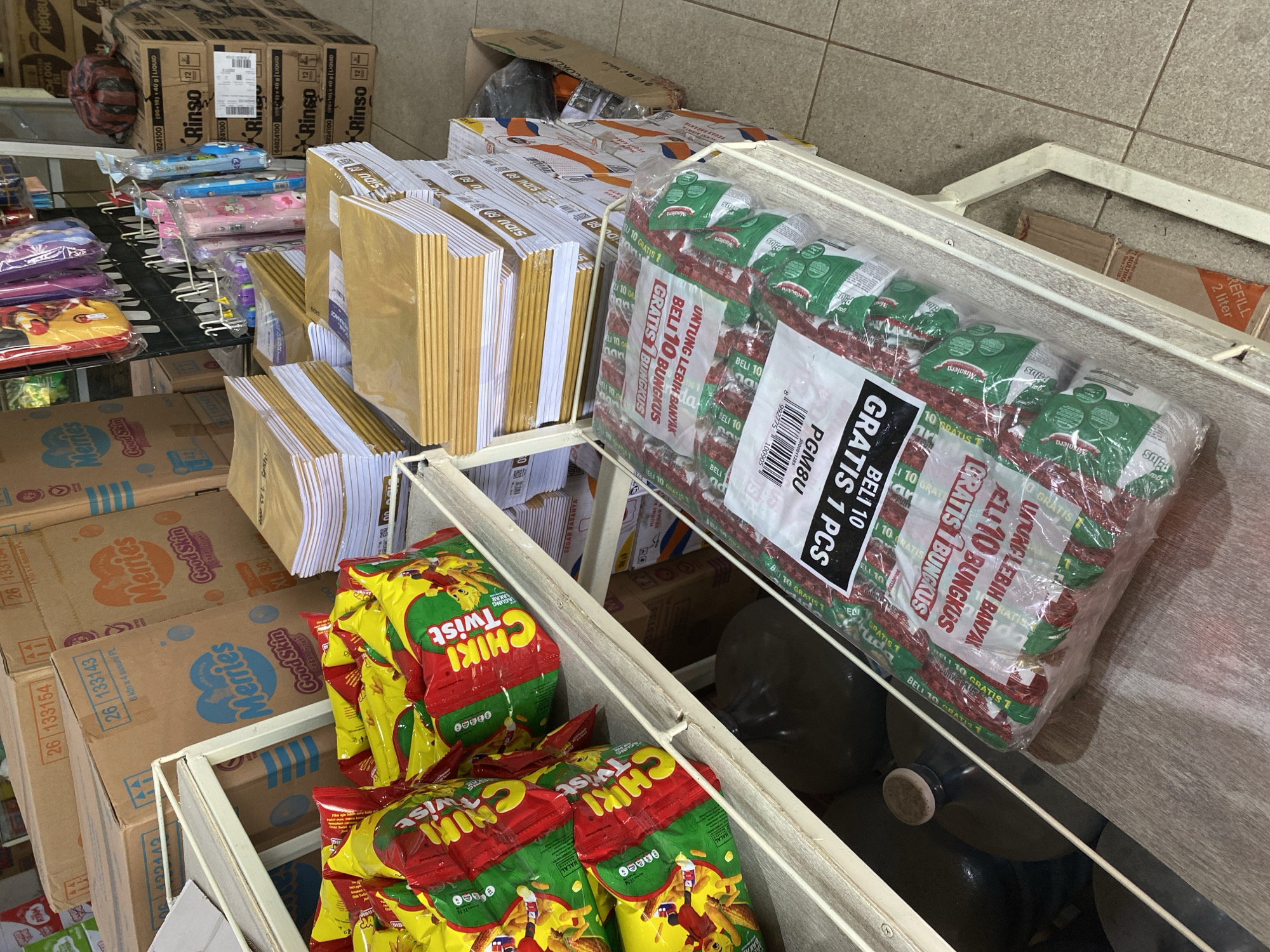 The image depicts a retail store aisle in Tanjong, Indonesia, stocked with various goods. On the left, shelves display bags of "Chiki Twist" snacks. To the right, stacked boxes of "Merries" diapers are visible, along with more boxes of diapers further in the background. In the foreground, a display of individually wrapped food items or snacks is packaged in clear plastic, featuring red and green designs and text in Indonesian, such as "GRATIS 10 BUNGKUS" (Free 10 packs) and "BELI 7 GRATIS 1 PCS" (Buy 7 Get 1 Free). Additional items like stationery or notebooks are also stacked on shelves, alongside hanging stationery items on a rack in the middle ground. The environment appears to be an indoor retail space with tiled flooring. No individuals are present in the scene.