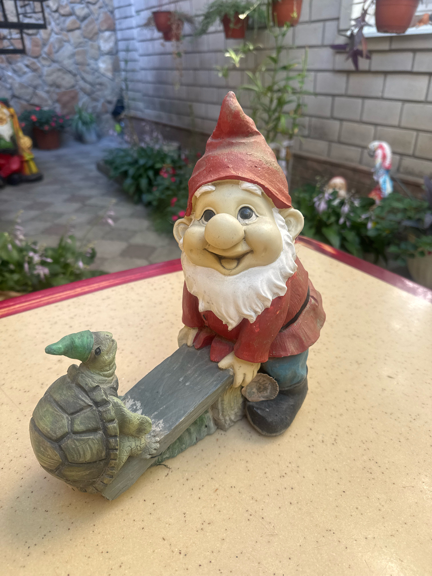 In the city of Anapa, Russia, a ceramic garden gnome and a ceramic turtle are positioned on a speckled yellow table. The gnome, dressed in a red tunic and hat, is leaning on a gray seesaw. The turtle, wearing a green pointed hat, is on the other end of the seesaw, facing left. In the background, a stone wall and a brick wall are visible, along with potted plants and other garden decorations. The overall setting appears to be an outdoor patio or garden area.