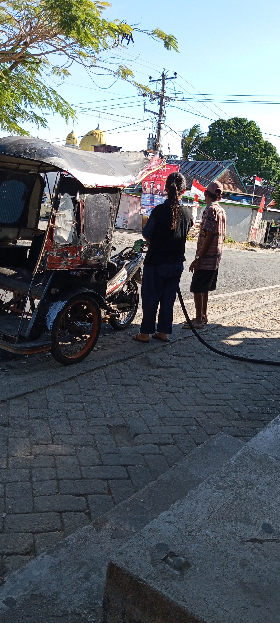 Two individuals, a woman wearing a dark blue long-sleeved shirt and dark pants, and a man in a plaid shirt and shorts, stand on a paved walkway in what appears to be an urban environment. The woman faces away from the camera, looking towards a three-wheeled vehicle that is parked next to her. The vehicle, which has a red and black color scheme and appears to be a modified motorcycle with a passenger compartment, is covered with a tattered black tarp. A black hose extends from the ground to the vehicle's engine area, suggesting it is being refueled or maintained. The man stands to the right of the woman, also facing away from the camera, looking in the same general direction. He is wearing a light-colored plaid shirt and dark shorts, and appears to be holding or interacting with the hose.

Behind the individuals and the vehicle, a street is visible, with buildings lining the opposite side. The buildings have a rustic appearance, with corrugated metal roofs and some displaying Indonesian flags. In the background, a building with a golden dome is visible, potentially indicating a religious structure. Electrical poles and wires crisscross the sky, typical of a developed urban area. The ground is paved with interlocking bricks, and a concrete step is visible in the foreground. The scene is illuminated by natural sunlight, casting shadows on the ground. The overall impression is a snapshot of everyday life in a moderately developed urban setting, possibly in Pakkanna, Indonesia, given the context provided.