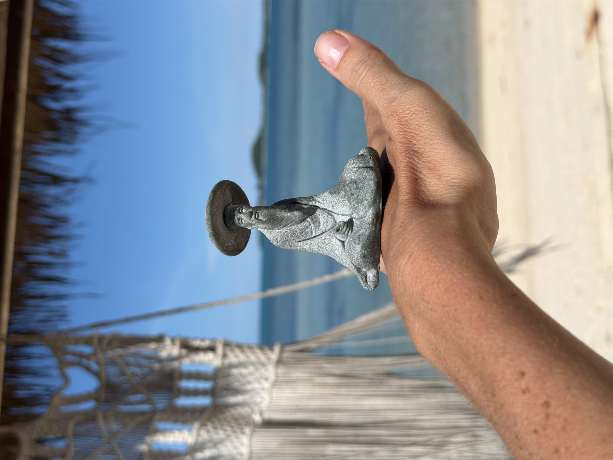 A person's arm and hand hold a small, grey statue of Jesus Christ. The statue is positioned in front of a blurred background of the ocean and beach. The image appears to be taken on a sunny day, with a clear blue sky visible. The presence of a hammock in the foreground suggests a relaxed, vacation-like setting. The shallow depth of field focuses attention on the statue and hand, with the background softly out of focus, creating a sense of peacefulness. There is no visible text in the image.
