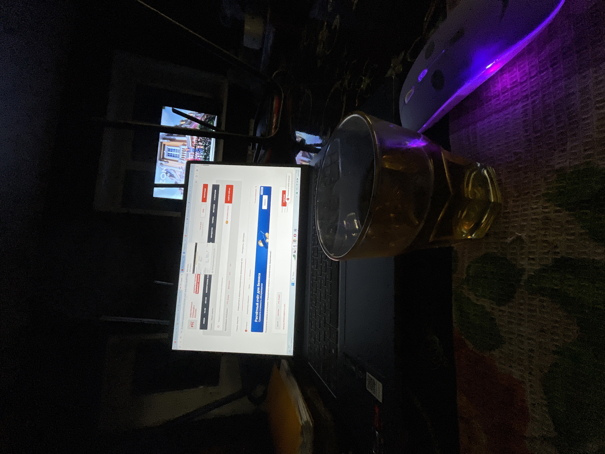 This image depicts a dimly lit indoor scene within the city of Chulym, Russia, observed during evening hours as indicated by the "19:34 МСК" (19:34 Moscow Standard Time) display. The primary subject is a silver-colored laptop, likely a Dell XPS, open and illuminated. Its screen displays a Russian-language website pertaining to an "ЭЛЕКТРОННАЯ ПЛОЩАДКА РОССИИ" (Electronic Platform of Russia), featuring the "PTC" logo and advertisements for services such as "Расчётный счёт для бизнеса" (Current account for business).
In the foreground, an amber-tinted glass tumbler containing a dark liquid is positioned on a patterned, woven surface. To the immediate right of the glass, a wireless computer mouse emits a distinct purple LED light. Further in the background, to the right of the laptop, a second illuminated screen or window displays an image of a building with a balcony, rendered in a bright, colorful style, potentially a European architectural scene. A partial text "ON" is visible on this background display. To the left of the laptop, a stack of papers or books is present on a dark surface. The overall environment suggests a personal workspace during nighttime. No individuals are visible in the scene.