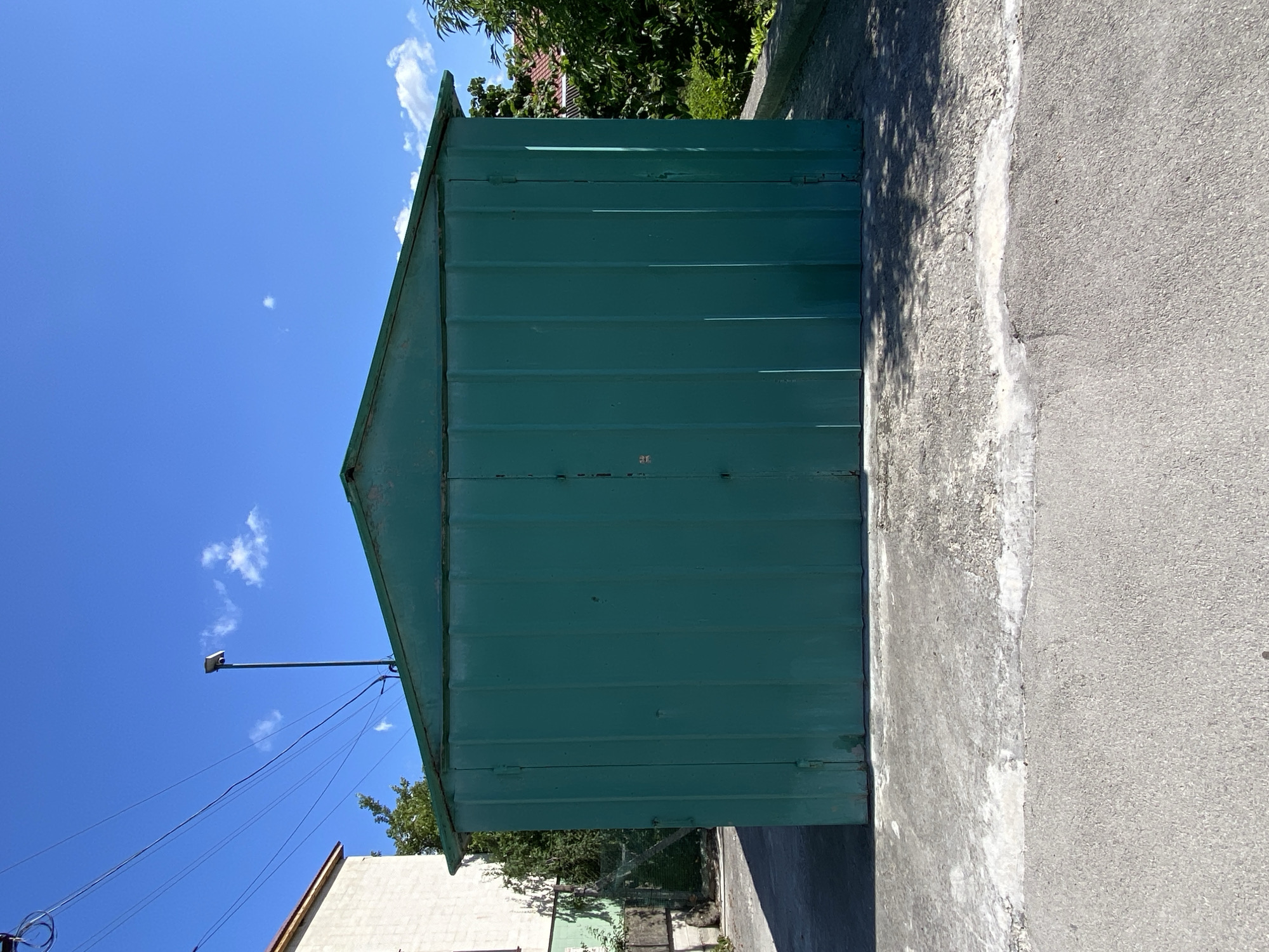 The image depicts a teal-colored corrugated metal garage situated outdoors under a clear blue sky with scattered white clouds. The garage has a peaked roof and double doors. In front of the garage, there is a concrete slab, which is cracked and weathered in places. Beyond the concrete slab, there is an asphalt surface. To the left of the garage, a portion of a light-colored brick building is visible, along with some greenery and a chain-link fence. To the right of the garage, trees and the roof of another building can be seen. A single light pole with a fixture at the top stands behind the garage, to the left of its center. Power lines are visible in the upper left quadrant of the image. The scene appears to be in a residential or semi-industrial area.
