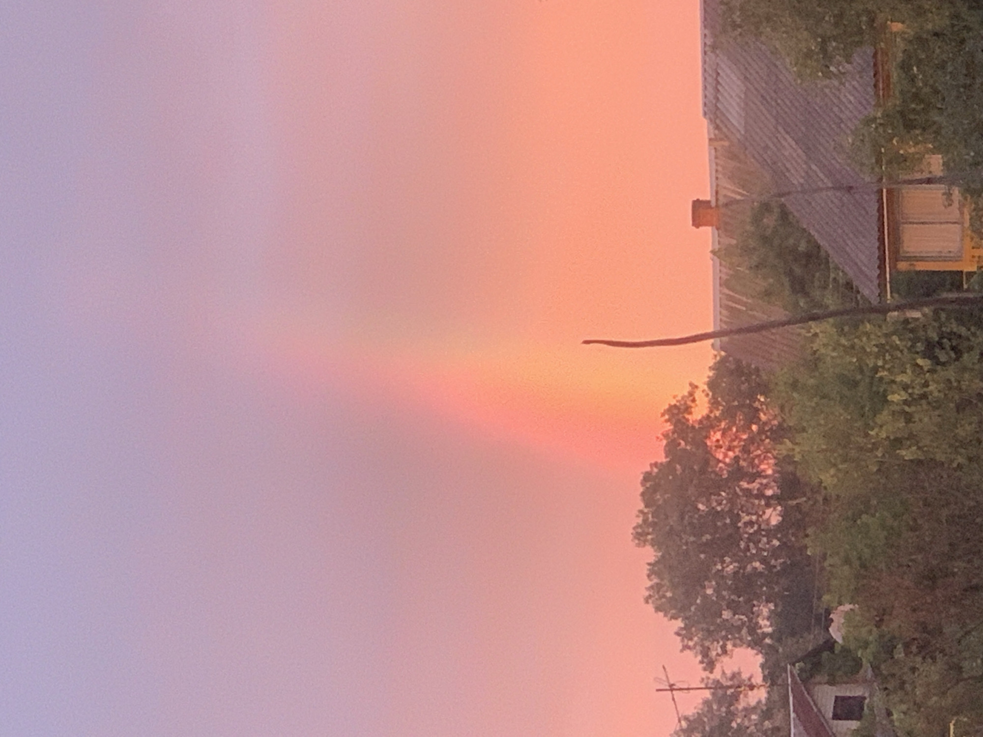 The image captures a vibrant sky with a rainbow arcing across it. The sky transitions from a soft purple at the top to a warm pink and orange hue towards the horizon. A faint rainbow, displaying shades of red and yellow, is visible against the colorful sky. Below the sky, the landscape features lush green trees and several buildings with tiled roofs. One building on the left has a visible window and a chimney on a structure behind it. A thin, dark vertical object, possibly a branch or a pole, extends upwards from the bottom center of the image, partially obscuring the view of the buildings and trees. The overall scene suggests a rural or suburban setting during either sunrise or sunset.