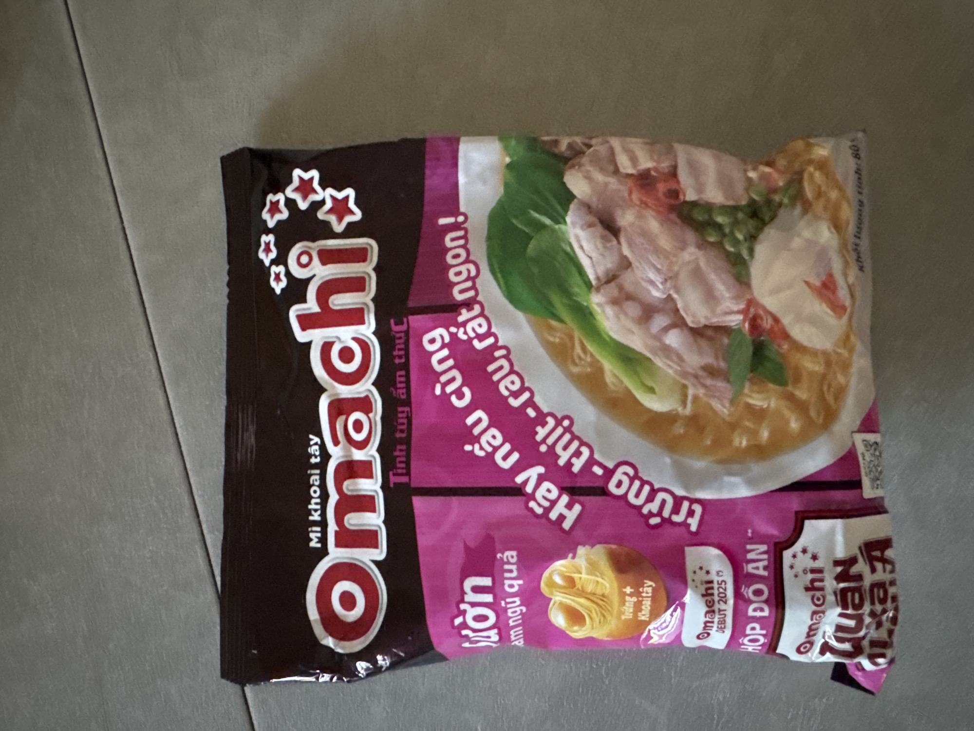 The image shows the front of a package of Omachi instant noodles. The package is primarily pink and brown and features a prominent red and white "Omachi" logo at the top. Below the logo, there are Vietnamese words. The lower portion of the package displays an illustration of the prepared noodles, featuring meat, vegetables, and broth. There is also text in Vietnamese on the lower left side of the package. The package is placed on a grey, textured surface, which appears to be a table or floor. The background is out of focus. There is no discernible presence of the city of Soloneshnoye, Russia, in this image.