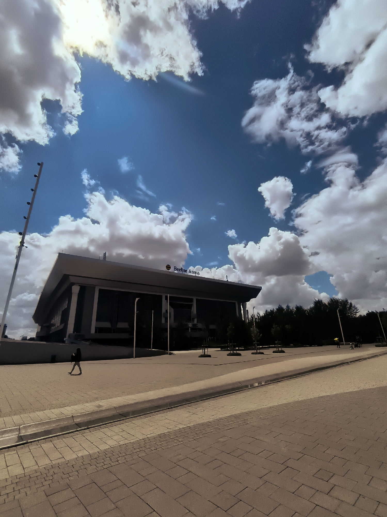 The image shows the Beeline Arena in Astana, Kazakhstan, on a cloudy day. The arena is a modern, multi-story building with a sleek, angular design. A large banner with the arena's name is visible on the roof. The sky is a vibrant blue, with large, fluffy white clouds casting shadows on the ground. A tall lamppost with multiple lights stands to the left of the arena. In the foreground, a person is walking on a paved walkway in front of the arena. Trees line the perimeter of the arena, and a few other people are visible in the distance. The overall scene depicts a public space in a city.