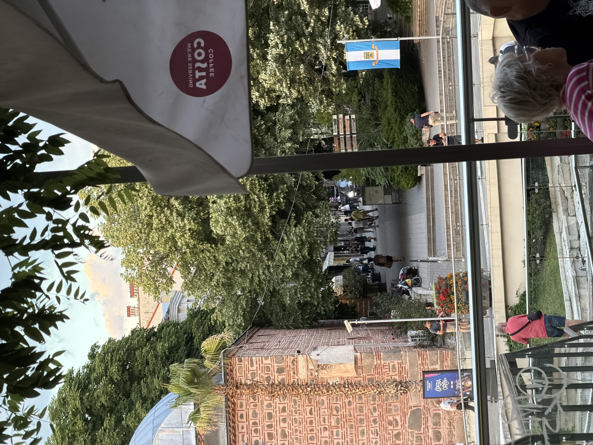 The image shows an outdoor view of a street in Plovdiv, Bulgaria, likely from a café or restaurant terrace. In the foreground, part of a Costa Coffee umbrella is visible. The street scene includes pedestrians walking and sitting at outdoor tables. A building with distinctive brickwork, possibly an old structure, is situated along the street. A flag with a blue and white emblem hangs above the street. People are seen on a higher terrace overlooking the street. The presence of street signs and outdoor seating areas indicates a public space.