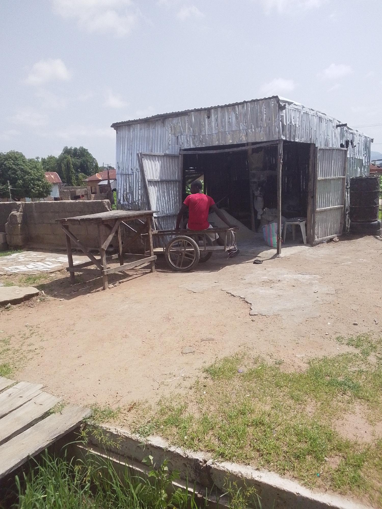 In a dusty outdoor setting, a man wearing a red shirt and white shorts sits on a wooden cart with two wheels. The cart is positioned in front of a corrugated metal structure that appears to be a shop or workshop, with its doors open to reveal the interior. To the left of the shop, a wooden table on legs stands on a paved area. The ground is a mix of dirt and patches of dry grass. In the background, there are trees and buildings with corrugated metal roofs. The sky is cloudy. The scene suggests a warm, possibly sunny day in a rural or semi-rural area. The presence of the cart and the open shop indicates some form of commerce or activity is taking place, although the specific nature of the activity is not clear from the image. The overall impression is one of a simple, functional environment.