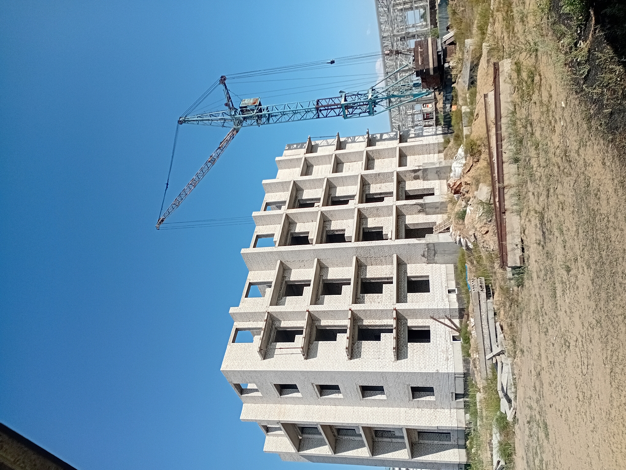 A multi-story building under construction, made of white brick, is shown against a clear blue sky. A large blue construction crane is positioned to the right of the building, with its boom extended over the structure. The ground in front of the building is rough and dusty, with piles of construction materials, including concrete slabs and rebar, scattered around. In the background, other construction elements, possibly parts of a larger structure, are visible. The scene appears to be a construction site in the city of Semey, Kazakhstan. There are no people visible in the image.