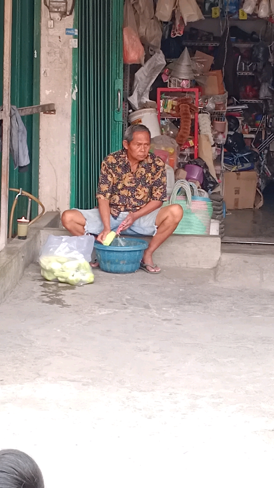 An elderly Indonesian man with short gray hair sits on concrete steps in front of a shop in Jambi, Indonesia. He is wearing a short-sleeved patterned shirt and denim shorts, with his legs spread apart. In his right hand, he holds a yellow object, possibly a sponge, which he is using to scrub something in a blue plastic basin placed on the step in front of him. A clear plastic bag filled with green produce, likely vegetables, sits to his left. Behind him, the shop displays a variety of items on shelves and racks, including plastic bags, containers, and tools, suggesting it is a general store or market stall. The shop's entrance is covered by a green, corrugated metal roll-up door. The ground in front of the steps is wet and appears to be a concrete walkway.