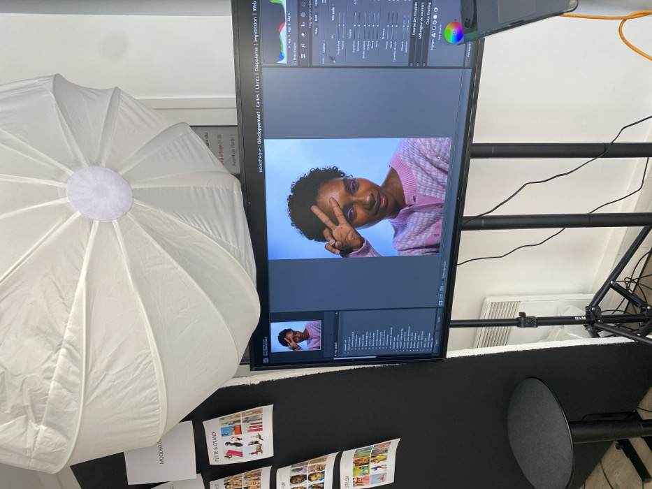 The image depicts an indoor scene within a photography studio or production office in Unknown, Unknown. A large, white, octagonal softbox light modifier is prominently positioned in the upper center. Below it, a large, flat-screen monitor displays a photo editing software interface, identified as Adobe Lightroom by the French text "Bibliothèque," "Développement," etc. The monitor's main panel shows a high-resolution portrait of a dark-skinned individual with short, dark, curly hair, wearing a pink and white checkered cardigan, striking a "peace" sign with their left hand near their face. The individual has purple eyeshadow, and the portrait background is solid light blue. A smaller thumbnail of the same image is visible on the left side of the screen, with various editing panels and a histogram on the right.
To the left of the monitor, a black partition or backdrop is visible, adorned with several printed papers. These papers include fashion mood board elements and text such as "MOODBOARD," "PETITE & GRANDE," and "STYLISM." A small, dark grey round stool with a black base is located in the lower-left corner next to this partition. The monitor rests on a black tripod stand. Numerous black cables are visible on the floor beneath and around the monitor. The background consists of a plain white wall, on which a white panel radiator is visible on the lower right. A piece of paper with French text, "Du chauffage ou de l'air conditionné (3 €)," is taped to the white wall above the radiator.