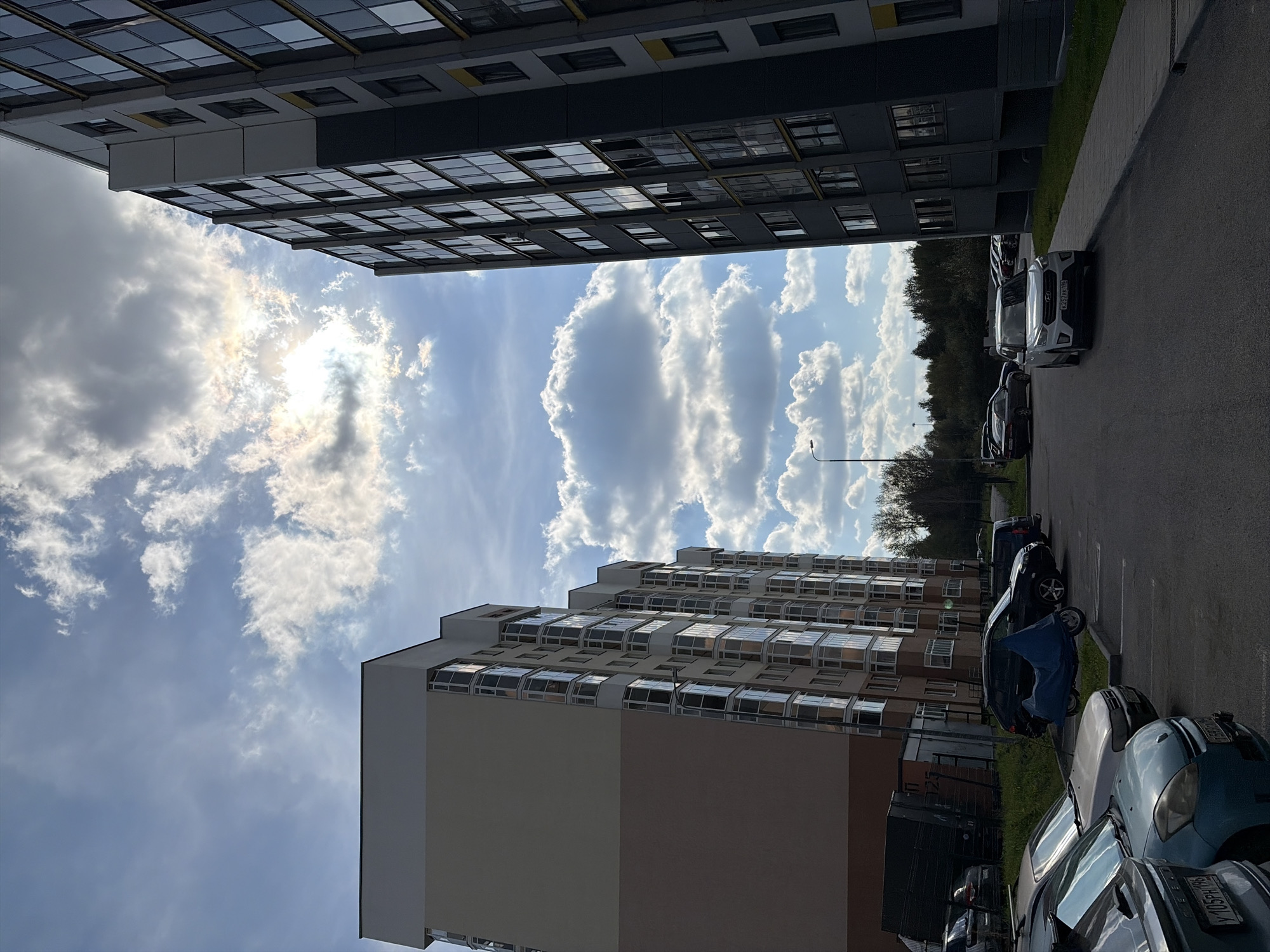 The image depicts a modern residential complex with two multi-story buildings flanking a parking lot. The buildings are constructed with a combination of light-colored facades and dark, reflective windows, suggesting a contemporary architectural style. In the foreground, a variety of cars are parked. One car, a white SUV, is prominently positioned near the right edge of the frame. A motorcycle covered with a blue tarp is also visible in the parking lot. The sky above is a mix of blue with scattered white clouds, and the sun is partially obscured, creating a dynamic lighting effect. The background includes trees and other residential structures, indicating an urban or suburban setting. The license plates visible on some of the vehicles, such as "Y 105PM 196" and "K457DK 196," suggest the location is in Russia. Based on the architectural style and visible license plates, this scene is likely located in the city of Yekaterinburg, Russia. There are no people clearly visible in the image.