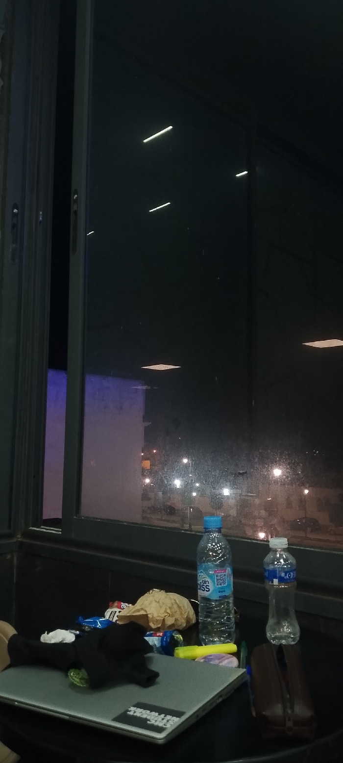 A cluttered desk sits before a large window at night. Two water bottles, snacks in a brown paper bag, and a dark object rest on a laptop. The laptop has a sticker. Outside, streetlights illuminate a cityscape. The window is dirty and reflects interior lights. The room is dimly lit, with a tall, purple-lit wall visible through the window frame.