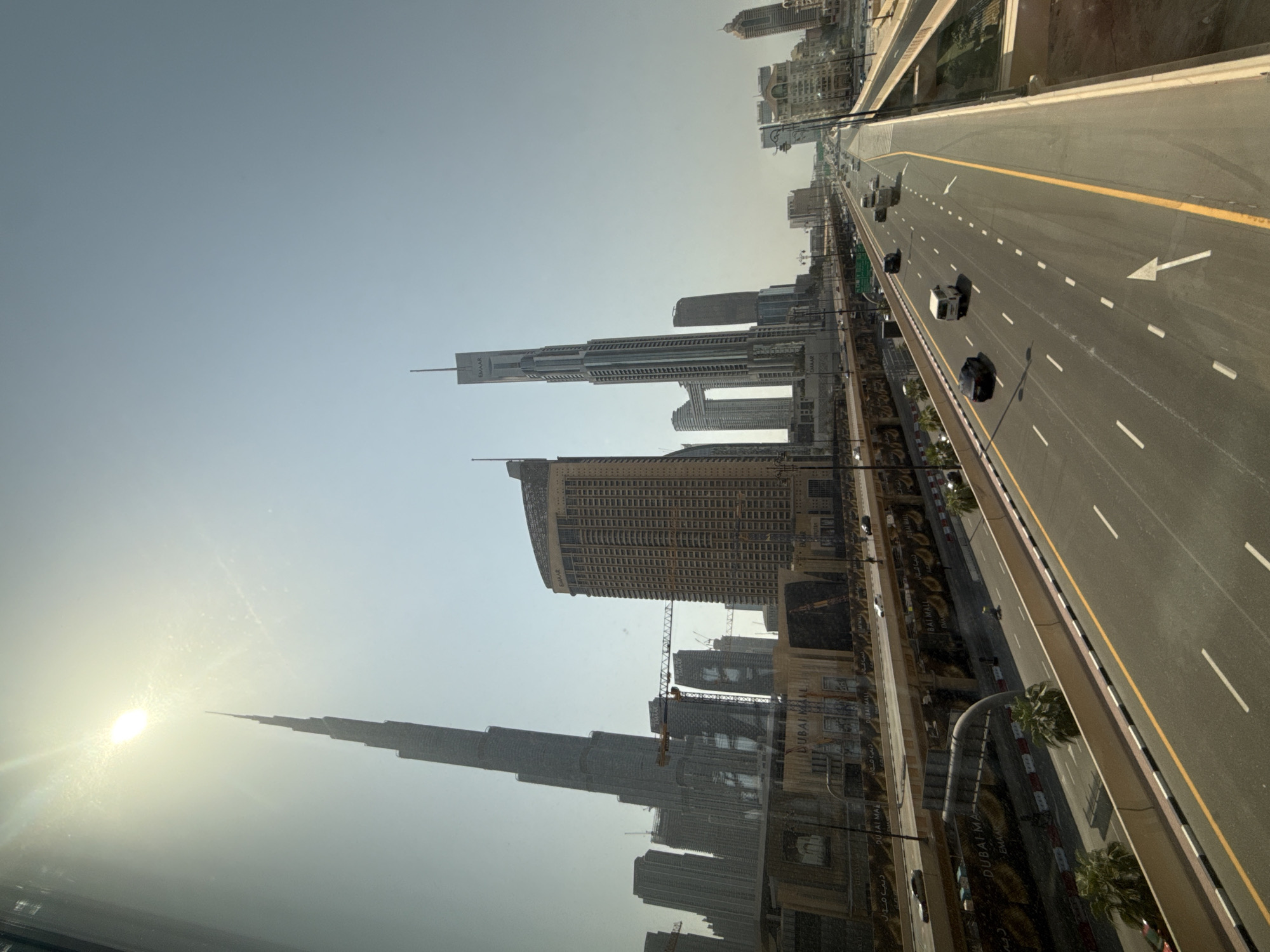 This image depicts a wide, elevated highway in Dubai, United Arab Emirates, with multiple lanes of traffic flowing in both directions. The highway is lined with concrete barriers and landscaping. In the background, the iconic Burj Khalifa dominates the skyline, alongside other modern skyscrapers. The sun is high in the sky, casting a bright light and creating some glare on the scene. Various vehicles, including cars and vans, are visible on the highway. The overall impression is of a busy, modern urban environment.