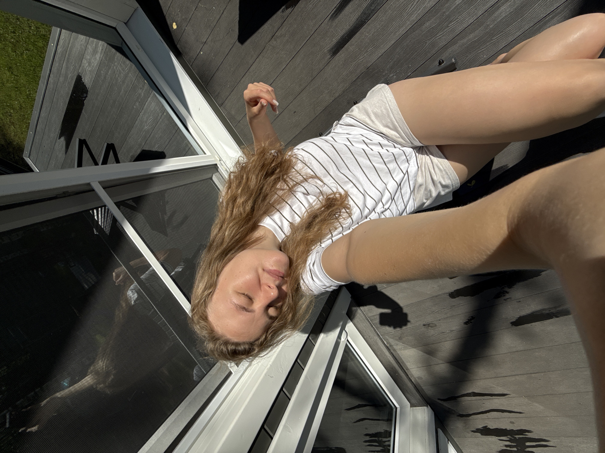 A young woman with long, wavy blonde hair is lying on her back on a dark wooden deck, with her eyes closed and her face tilted towards the sun. She is wearing a white t-shirt with thin black stripes and light-colored shorts. Her left arm is extended, holding what appears to be a phone, suggesting she is taking a selfie. The deck is next to a large sliding glass door with white framing. Shadows are cast on the deck, indicating a sunny day. In the background, through the glass, a glimpse of green grass and possibly trees is visible. The overall scene depicts a relaxed moment outdoors.