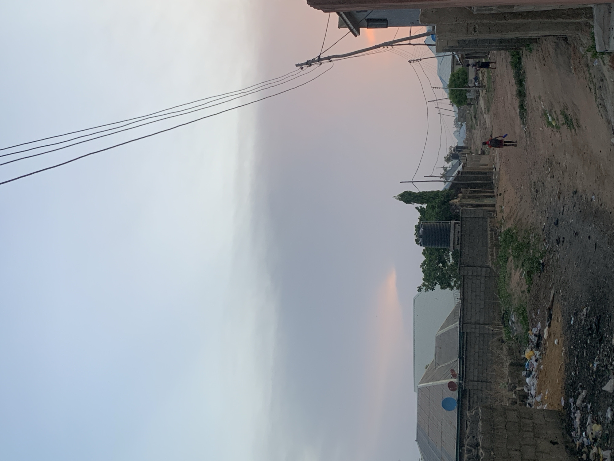 The image depicts a dirt road in Lokoja, Nigeria, lined with simple concrete-block structures. Overhead power lines stretch across the scene. Litter is visible on the sides of the road. Two individuals are walking on the road: one is in the distance while another is closer wearing a red shirt and carrying objects. A water tank and some trees are present behind the wall. The sky is overcast.