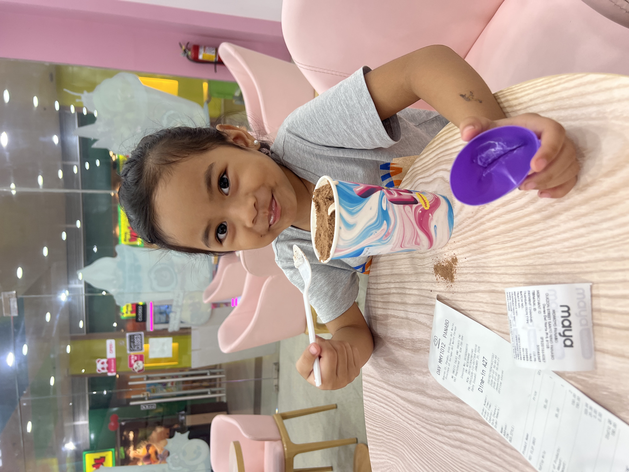 A young Filipino girl with dark hair tied back in a ponytail sits at a light-colored wooden table in what appears to be an indoor establishment. She is wearing a grey t-shirt and has a small, faint tattoo on her left forearm. She is holding a white spoon with a small amount of brown ice cream or dessert on it, and in her right hand, she holds a small purple plastic cup. In front of her is a paper cup with a colorful swirl design containing a large scoop of brown ice cream. A receipt from "MOMOYO TOYS AND TREATS INC." in Panabo City, Davao del Norte, Philippines is partially visible on the table, along with a smaller receipt from "maya". The background shows a glass storefront with various signs and a glimpse of pink furniture, suggesting a commercial or dining setting. The overall lighting is bright and appears to be artificial.