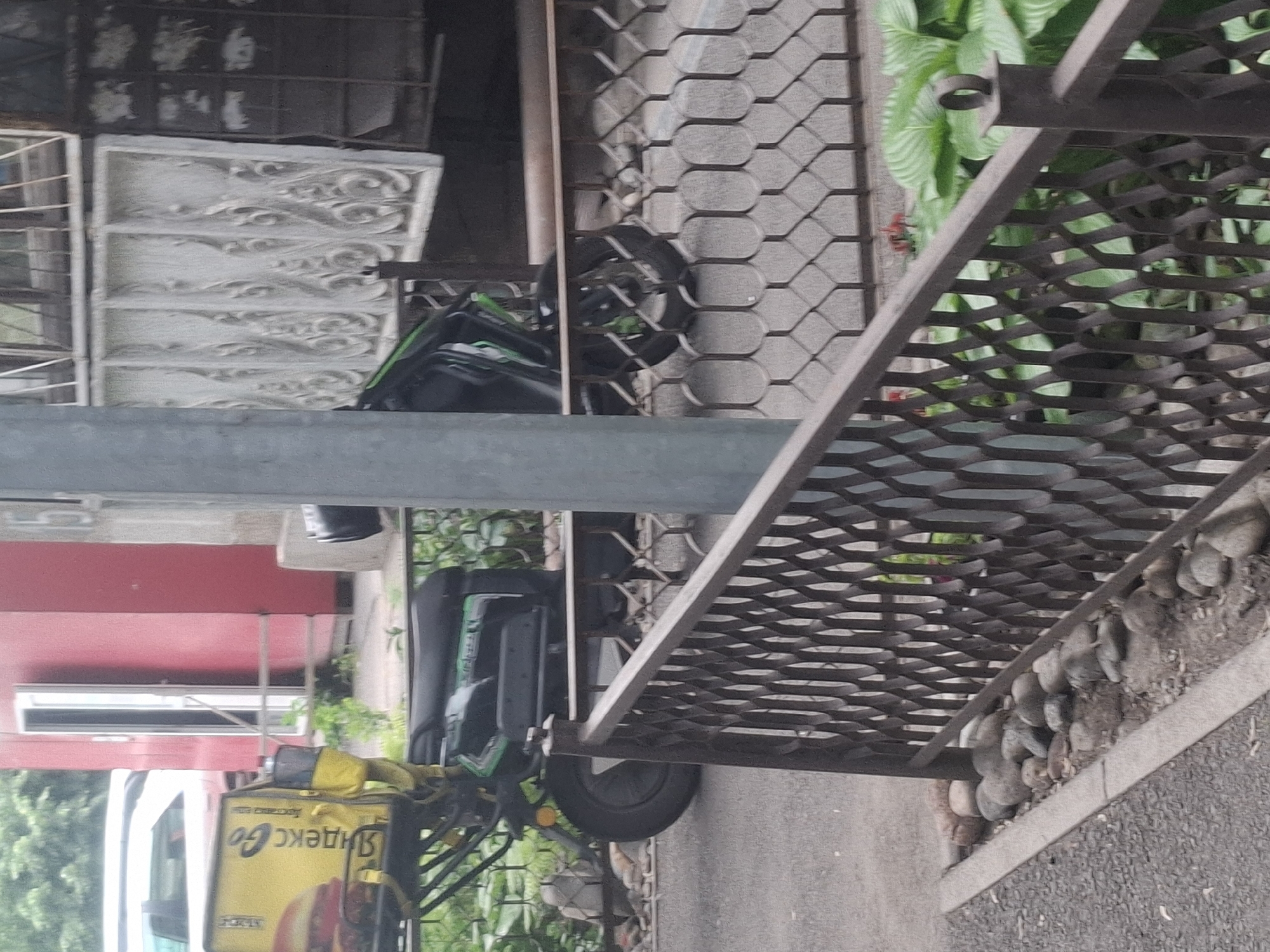 The image shows a delivery scooter with a yellow Yandex.Go insulated bag parked on a sidewalk next to a building with a decorative metal grate. The scooter is black with green accents and has a large rear cargo area. A white car is partially visible in the background, along with a red building and some greenery. The ground is paved and there are rocks and plants visible along the side of the building. The scene appears to be in an urban environment, likely a residential or commercial area.
