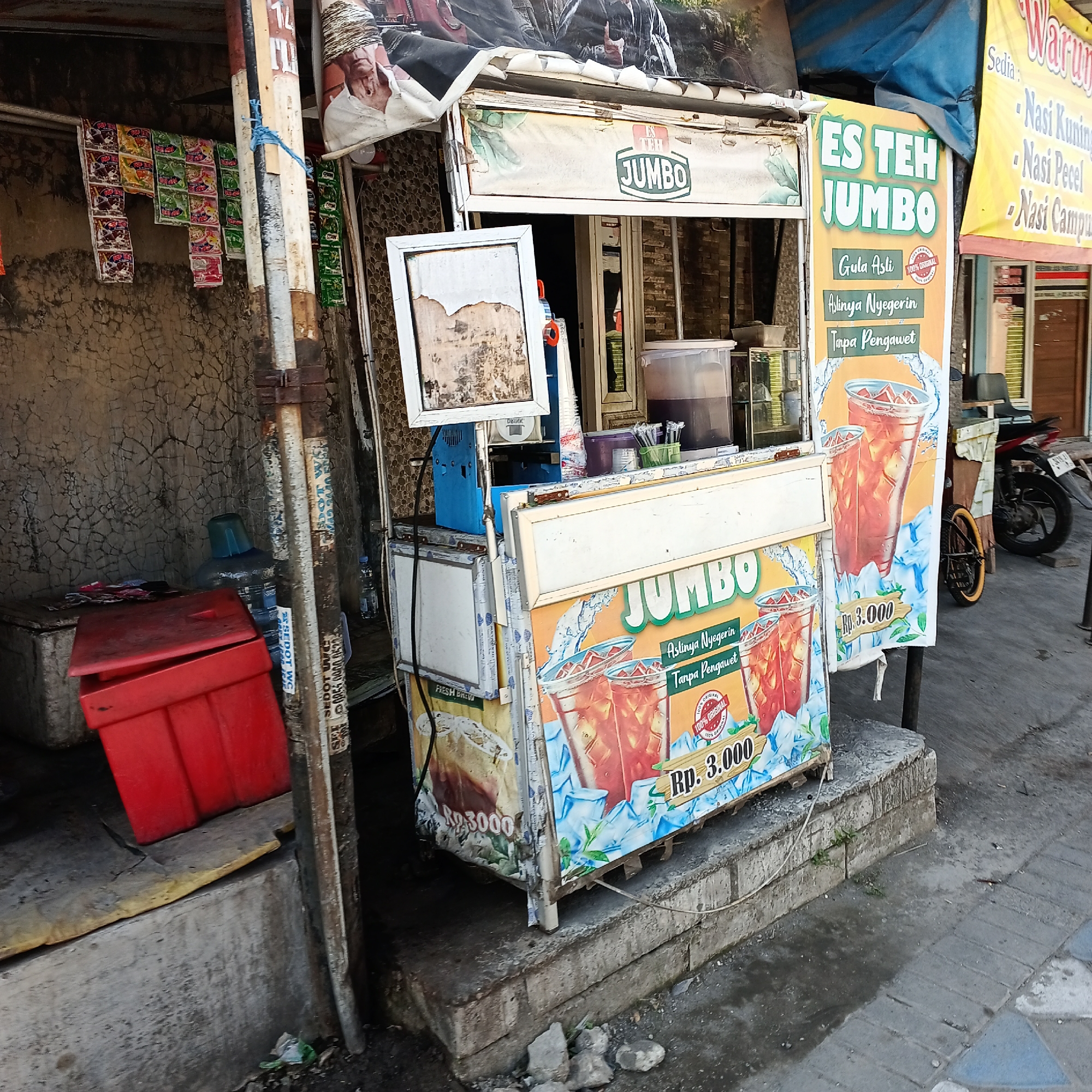 A street-level view captures a food and beverage stall in Kepuh Kiriman, Indonesia. The stall displays an awning with the text "ES TEH JUMBO" and features colorful graphics of iced tea. Various beverages are visible within the stall, including large dispensers of dark liquid and bottled water. Next to the stall, a red cooler box sits on a concrete platform. A motorcycle is parked nearby, partially obscuring some of the stall's signage and the surrounding environment. The area appears to be an outdoor, urban setting with a concrete sidewalk and cracked, textured walls. Packaging for snack items is hanging on the wall to the left of the stall.