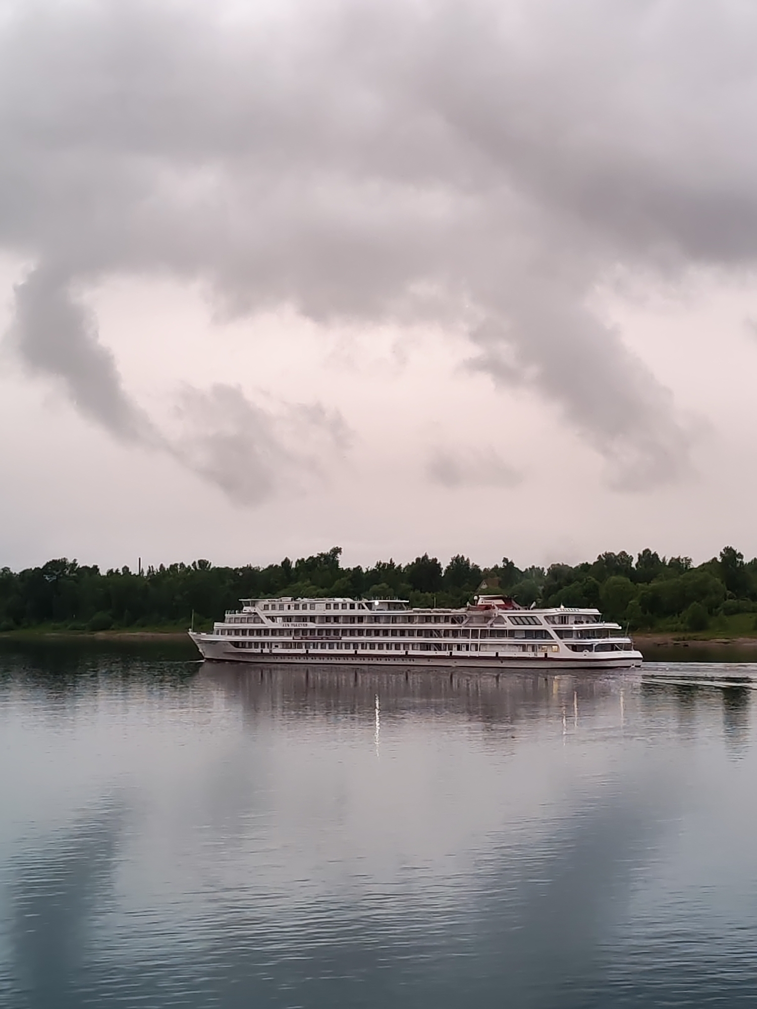 The image depicts a river scene in Bol'shie Mkhi, Russia, under a cloudy sky. A white cruise ship with multiple decks is moving along the water, its reflection visible on the river's surface. The river is flanked by a lush green shoreline of trees and vegetation. A tall smokestack rises in the background behind the trees on the far left. The overcast sky contributes to a muted color palette throughout the scene.