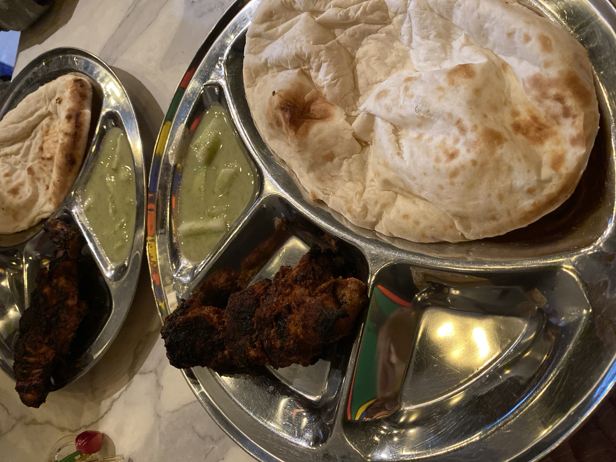 The image shows two compartmentalized metal trays of food, likely taken in a restaurant in Dhaka, Bangladesh, given the style of food. The tray on the right holds a large piece of flatbread, a portion of green chutney or sauce, and several pieces of tandoori chicken. One compartment is empty. The tray on the left contains a smaller piece of flatbread, a similar green chutney or sauce, and several pieces of tandoori chicken. Both trays are placed on a marbled surface, possibly a table. A small artificial flower can be seen in the bottom left of the image.