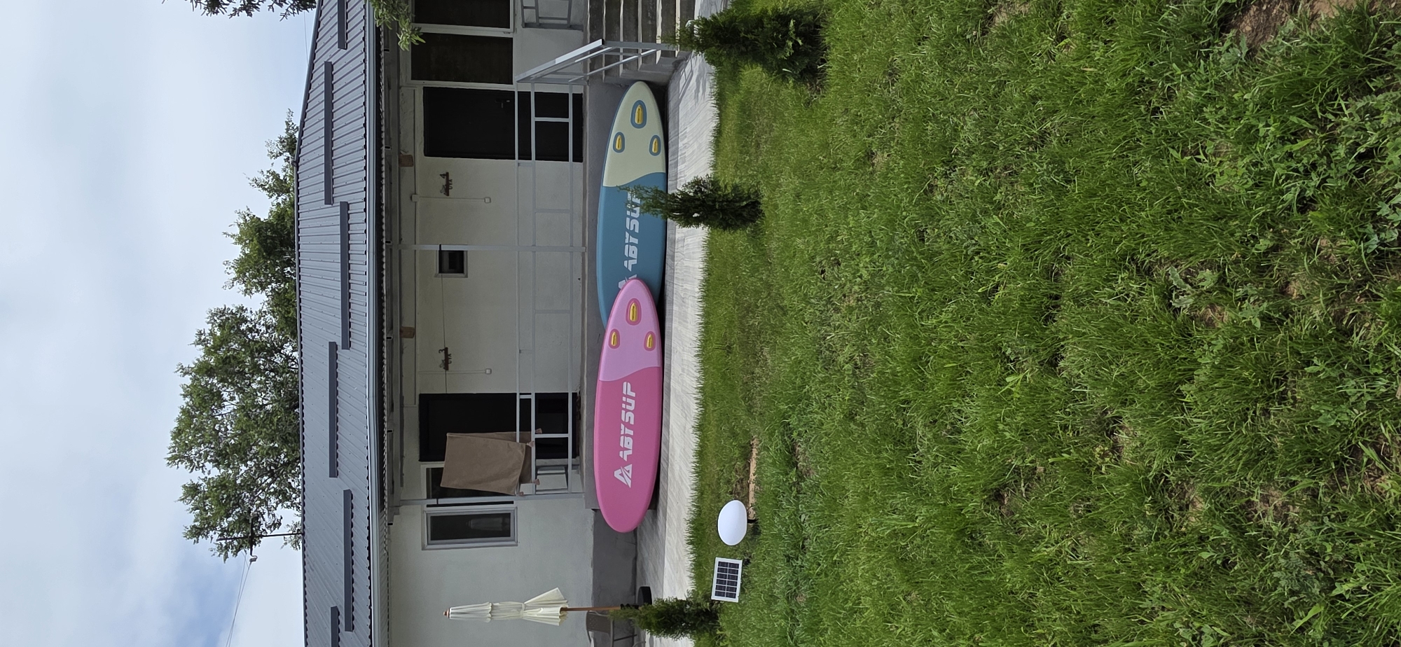 An outdoor shot shows a building with a gray metal roof and white siding. Two inflatable paddleboards are leaning against the building on a paved area. The pink paddleboard is on the left, and a blue and yellow paddleboard is on the right. There is a white outdoor umbrella to the left of the paddleboards. In front of the building is a grassy area with two small evergreen trees, a solar-powered light, and a solar panel. The sky is cloudy. The location is unknown.