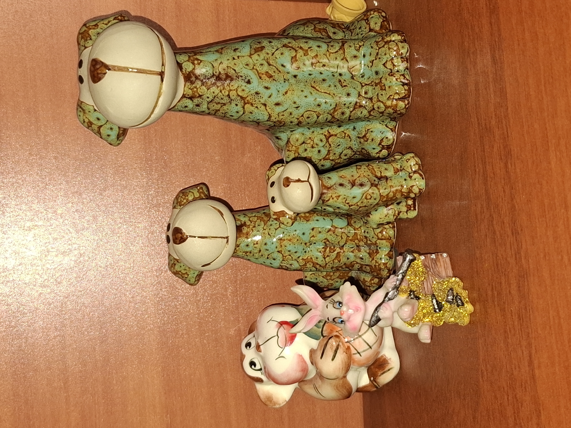 The image shows a collection of ceramic figurines on a wooden shelf against a textured brown wall. The figurines include two large dogs with green and brown speckle glaze, and one smaller dog of similar design. To the left of the dogs is a ceramic monkey-like figure holding a red flower. In front of the monkey is a small pink bunny figurine holding a broom with a yellow sparkly attachment. The overall scene is an indoor display of decorative items. The location in Almaty, Kazakhstan, is not specified.
