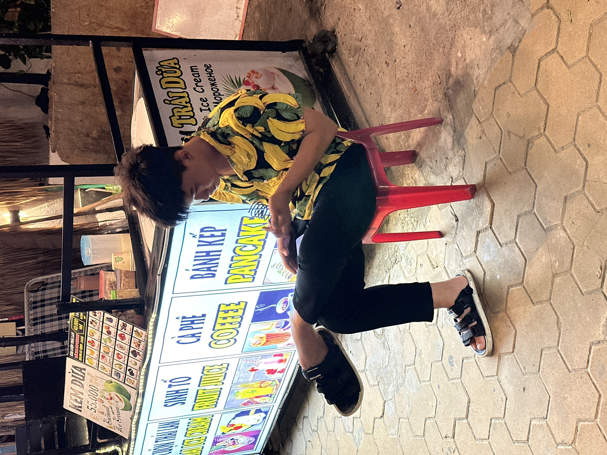 A young Vietnamese male with dark hair is seated on a red plastic stool in front of a food stall. He is wearing a yellow short-sleeved shirt with a banana print and black pants. His left leg is crossed over his right, and he is looking down at his phone, which he holds in his hands. The food stall has a white illuminated sign with text in Vietnamese and English, advertising various food and drink items such as "KEM DUA" (Coconut Ice Cream), "SINH TO FRUIT JUICE" (Fruit Juice), "CA PHE COFFEE" (Coffee), and "BANH KEP PANCAKE" (Pancake). There are pictures of the food items on the sign. The stall is located on a sidewalk paved with hexagonal tiles. In the background, there is a checkered cloth draped over something, and some greenery. The lighting suggests it is nighttime or a dimly lit area. The scene appears to be set in Phu Thuy Ward, Vietnam, as indicated by the Vietnamese text on the sign.