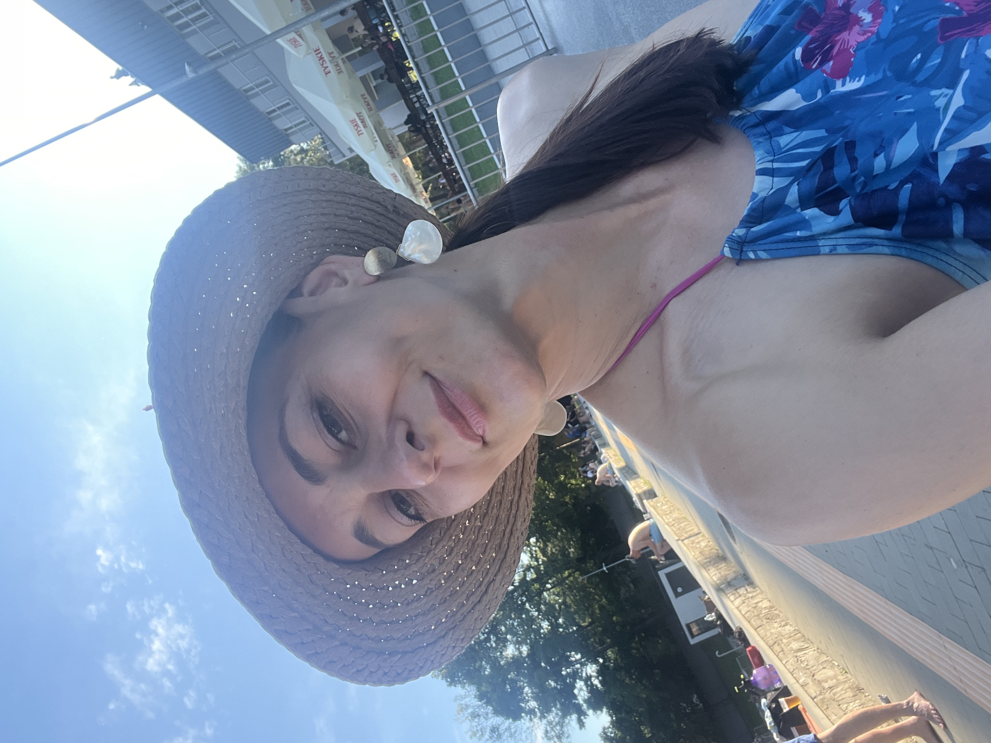 This is a selfie of a woman wearing a straw hat and a blue floral swimsuit. She is outdoors, with a bright blue sky and scattered white clouds in the background. Sunlight suggests it is daytime, likely sunny.
The woman is looking towards the camera with a slight smile. She is wearing a pair of circular, possibly shell-like earrings. Her hair is dark brown and long, falling over her shoulder. The swimsuit has a halter neck with pink straps.
In the background, there are hints of a setting that could be a resort or outdoor venue. A building with several windows is visible, and a white tent with text is partially obscured. Other people are faintly visible in the distance, some appearing to be seated or standing. The ground in the foreground is paved, with a stone wall or border.
The overall impression is one of a casual, relaxed moment, possibly during a vacation or a day out in the sun. The angle of the photograph and the lighting indicate it was taken during daylight hours.