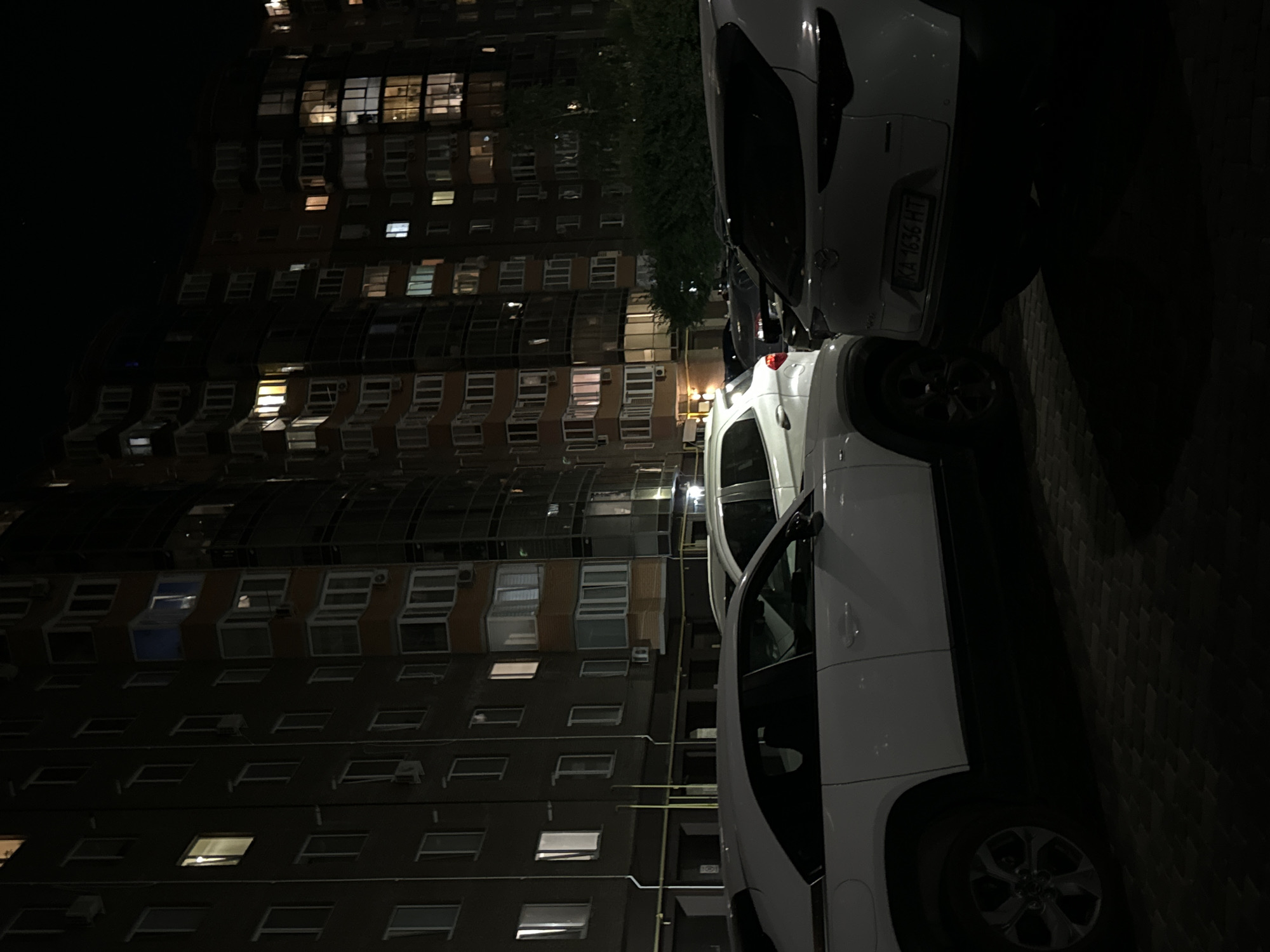 The image depicts a nighttime scene in Vinnytsia, Ukraine, featuring a modern apartment building with illuminated windows. In the foreground, several white cars are parked on a paved surface. The building's facade displays a mix of dark and orange sections, with numerous windows showing interior lights, suggesting occupancy. The overall atmosphere is one of a residential urban environment after dark.