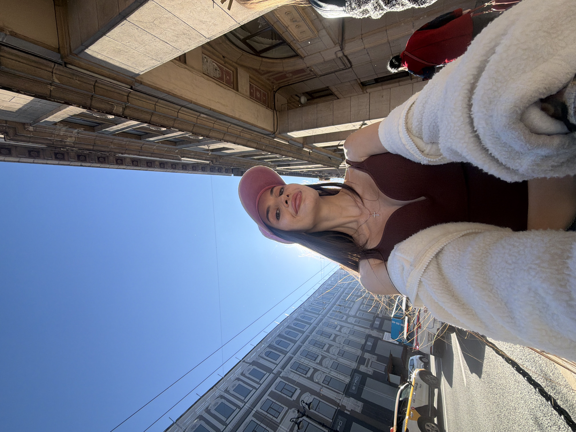 The image shows a woman taking a selfie on a sunny day in a city. She is wearing a maroon v-neck top, a cream-colored fuzzy jacket, and a pink baseball cap. She has long dark hair and is wearing a cross necklace. In the background, there are buildings with classical architecture, some with ornate detailing. A street with cars, including a yellow and white taxi, is visible, suggesting an urban environment. The sky is clear and blue. The architectural style of the buildings and the presence of a taxi that appears to be a Yandex taxi are indicative of Saint Petersburg, Russia.